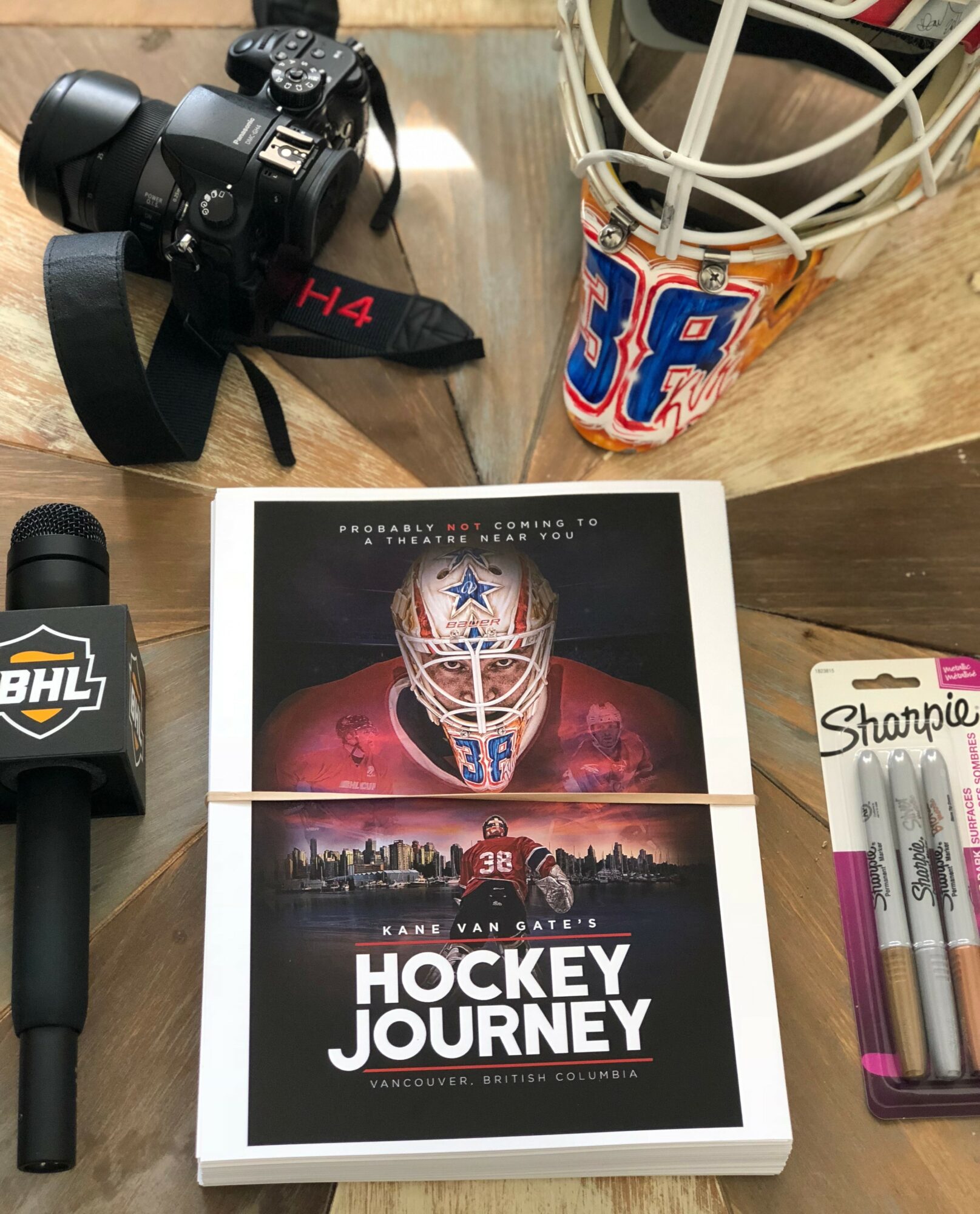 Camera, hockey book titled 'Hockey Journey' with a Dallas Cowboys helmet, Sharpie pens, and a drink on a wooden table.