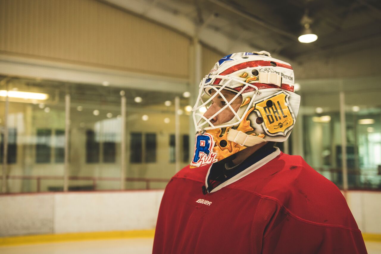Ice hockey goalie in red jersey wearing helmet and mask, standing in rink, facing right, with glass and ice in background.