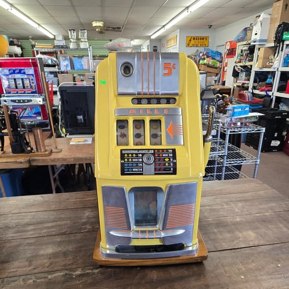 Vintage slot machine with yellow and gray colors on a wooden table in a store, surrounded by shelves and equipment.
