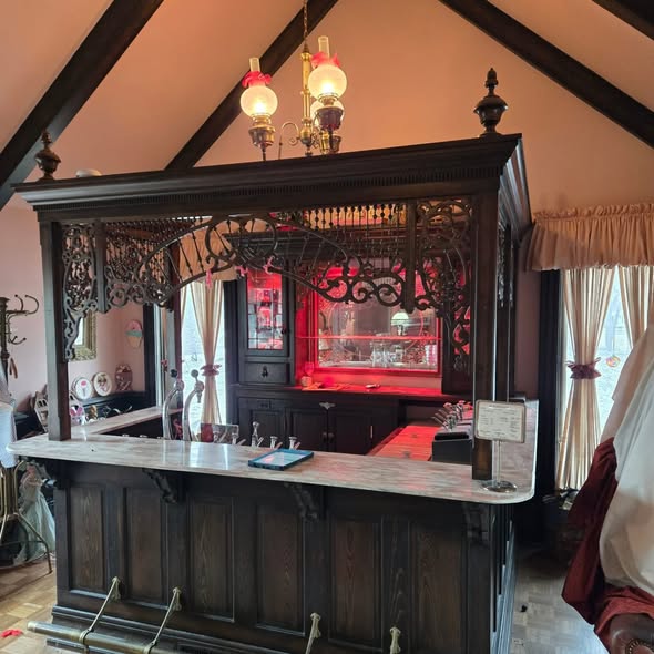 Dark wooden bar with ornate carvings, marble countertop, and hanging lights, in a room with high ceiling and curtains.