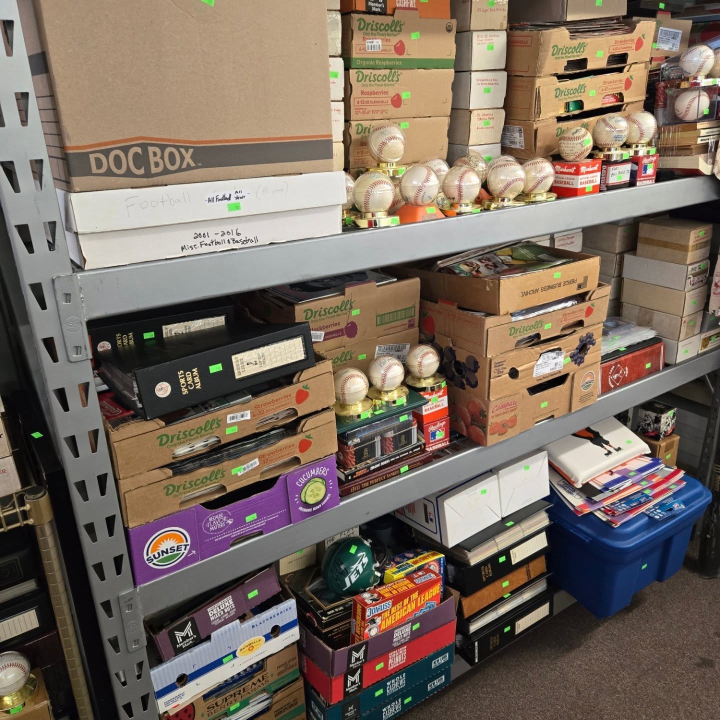 Shelves stocked with boxes of baseballs, some in cardboard boxes and others loose, with a blue storage bin on the floor.