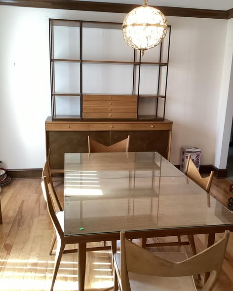 Dining room with a glass table, six wooden chairs, a wooden sideboard, and a large empty bookshelf, illuminated by a hanging light fixture.
