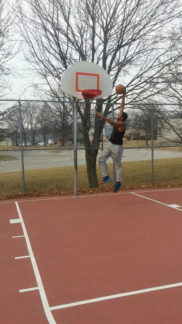Person jumping to shoot basketball on outdoor court with trees and fence in background.