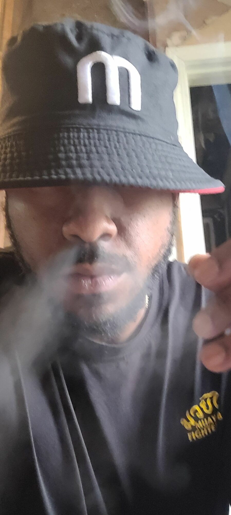 Person wearing a black cap and jacket, exhaling smoke, with a blurred background.