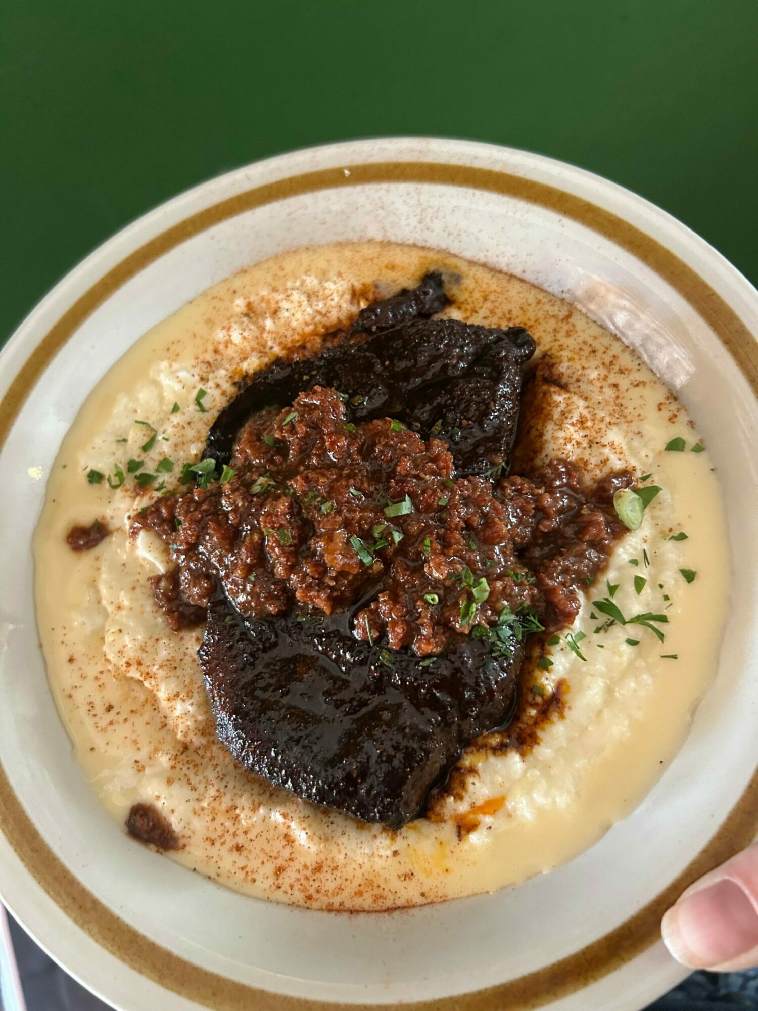Bowl of creamy porridge topped with dark sauce, meat, and chopped herbs, with a hand holding the bowl's edge.
