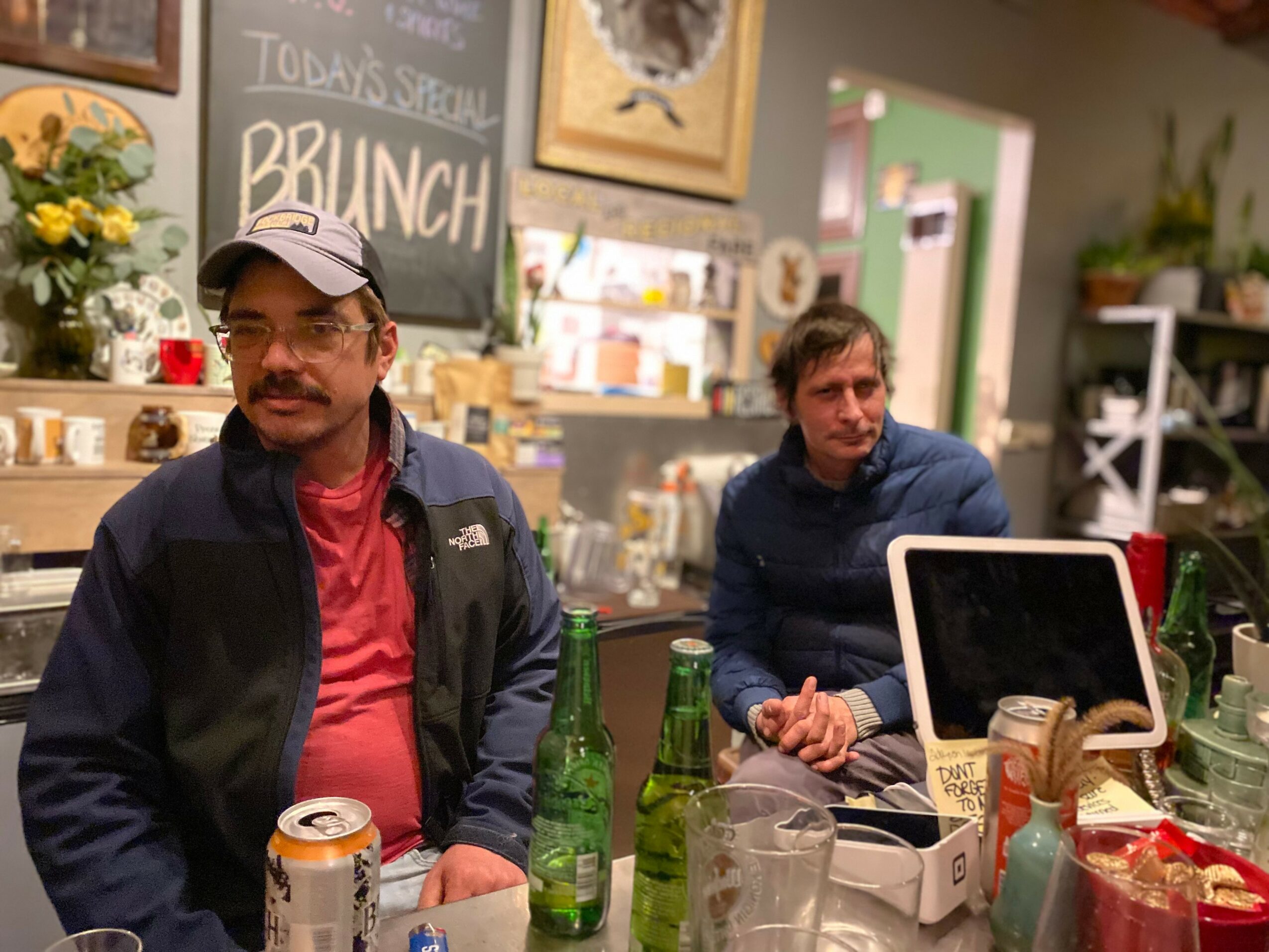 Two men sitting at a table in a cozy cafe, with a chalkboard menu and decorative plants in the background.