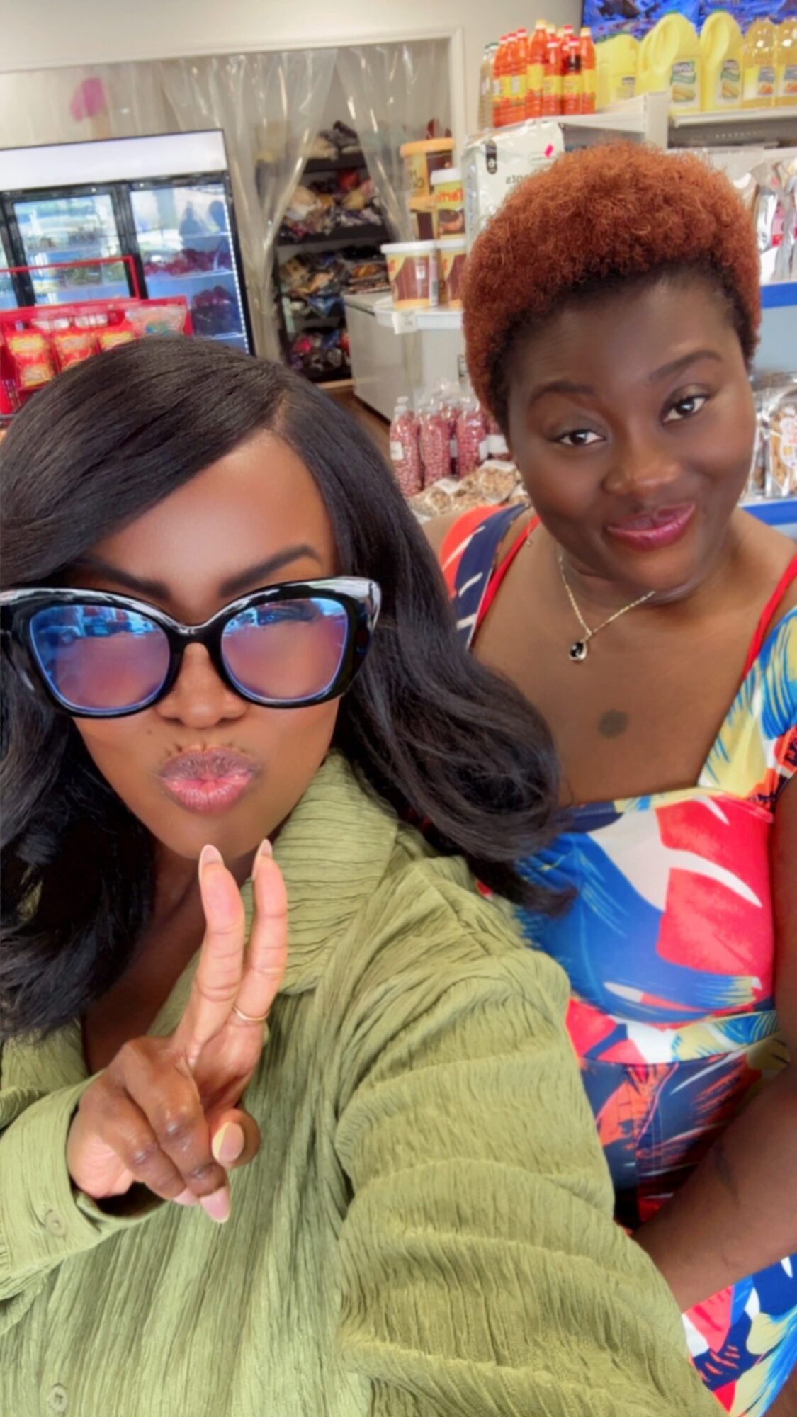 Two women posing in a store, one wearing large glasses and making a peace sign, the other smiling with short curly hair.