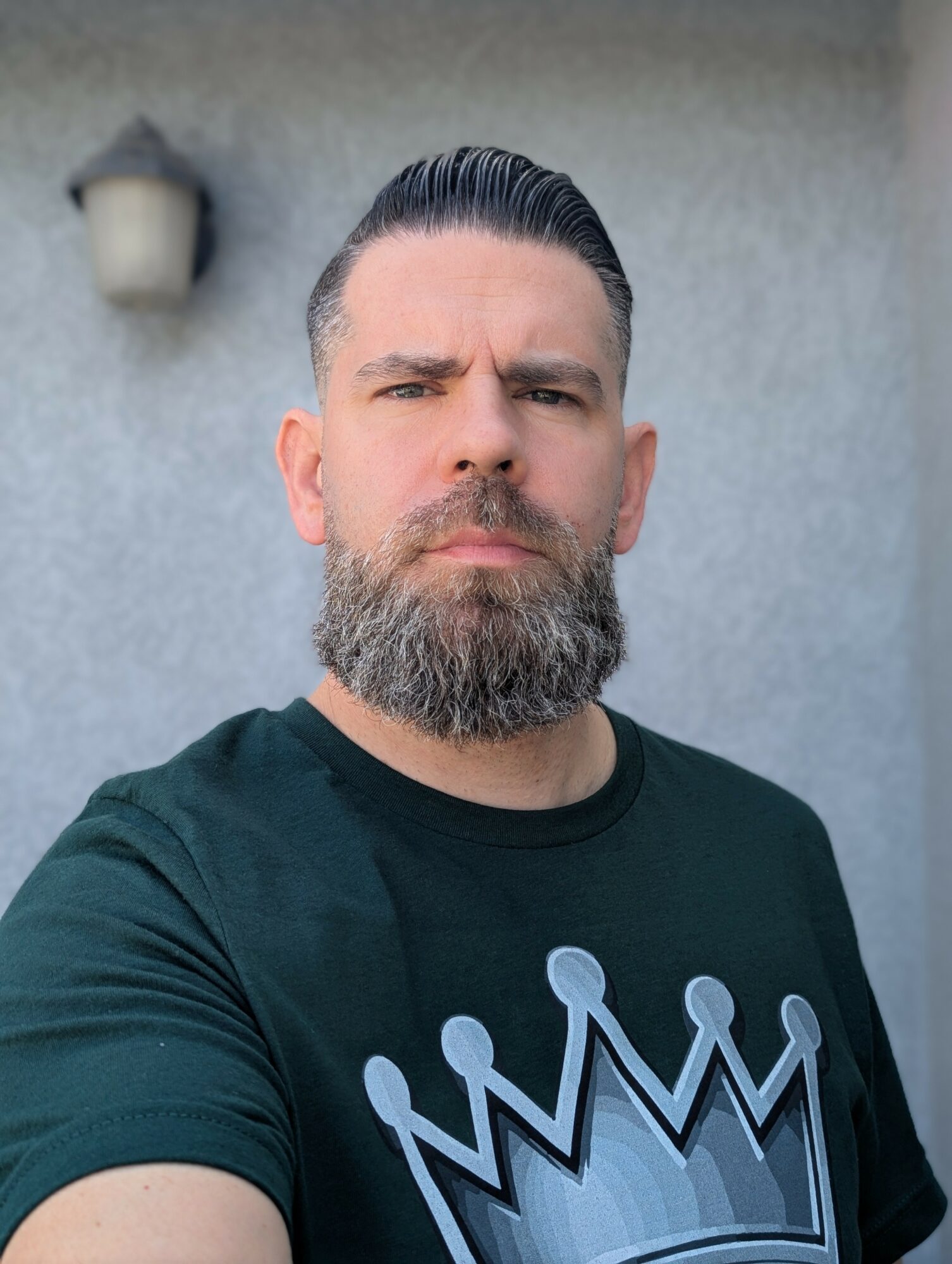 Man with a beard and slicked-back hair wearing a black T-shirt with a crown graphic, standing outdoors against a gray wall.