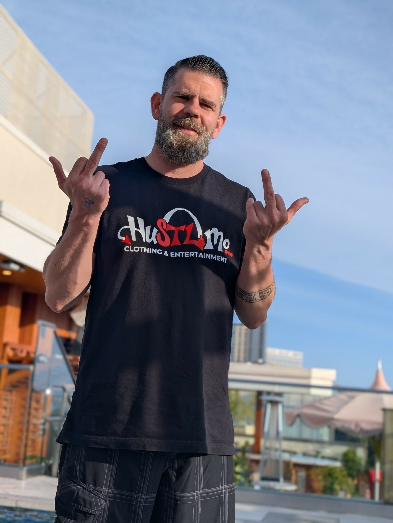 Man with beard and short hair making hand gestures outdoors, wearing a black T-shirt with red and white text, smiling.