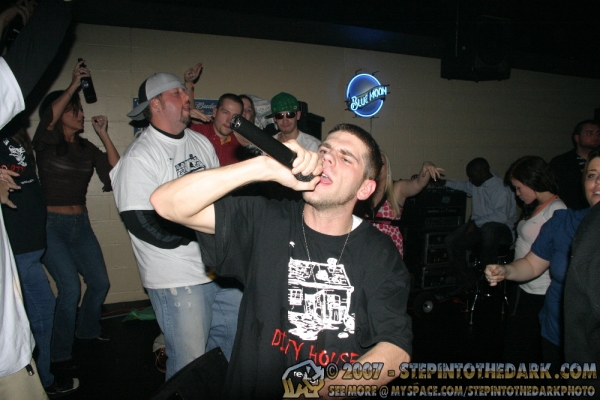Young man singing into a microphone at a party with people dancing in the background.