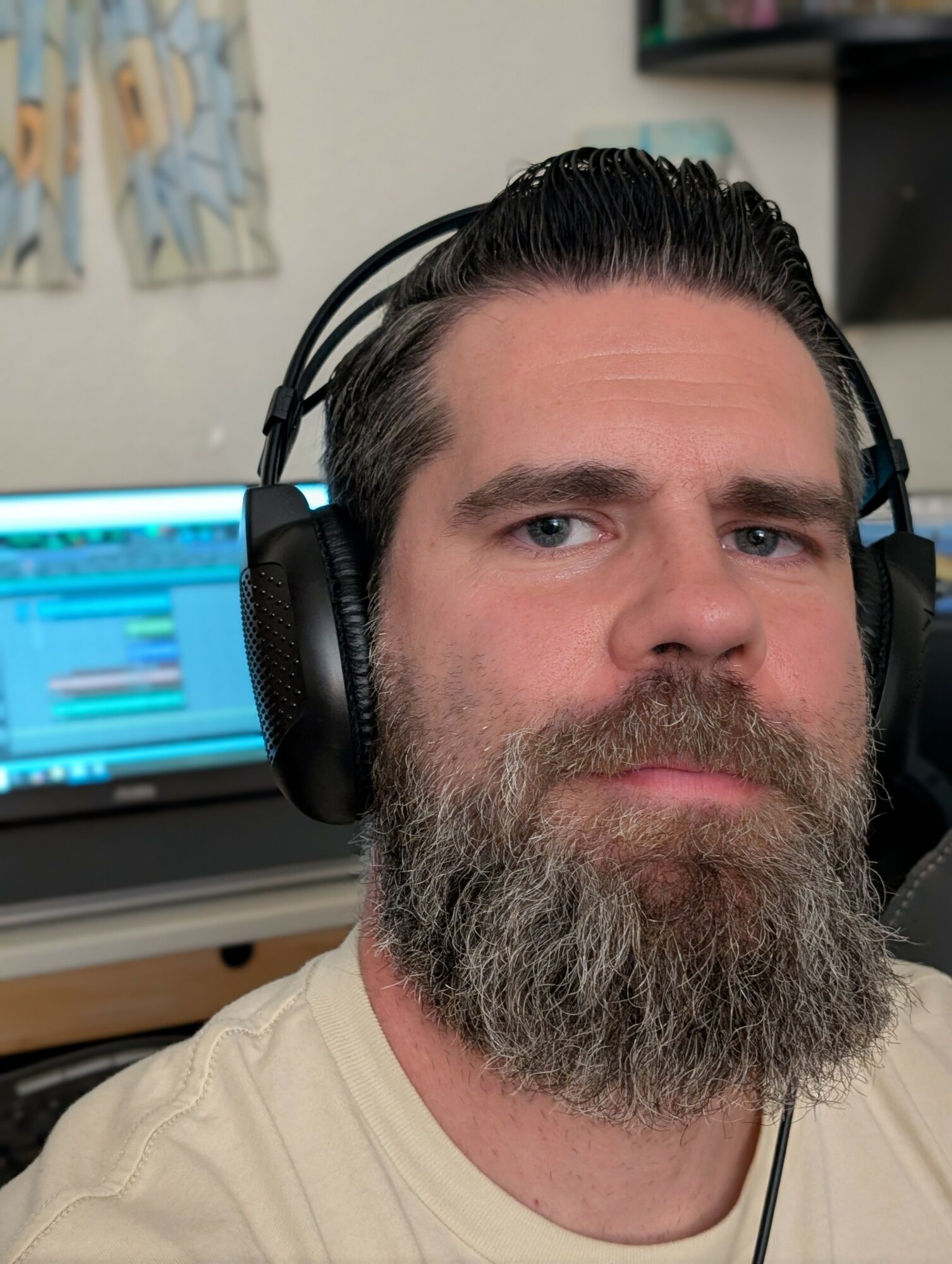 Man with dark hair and beard wearing headphones, in front of a computer screen, in a room with wall art.