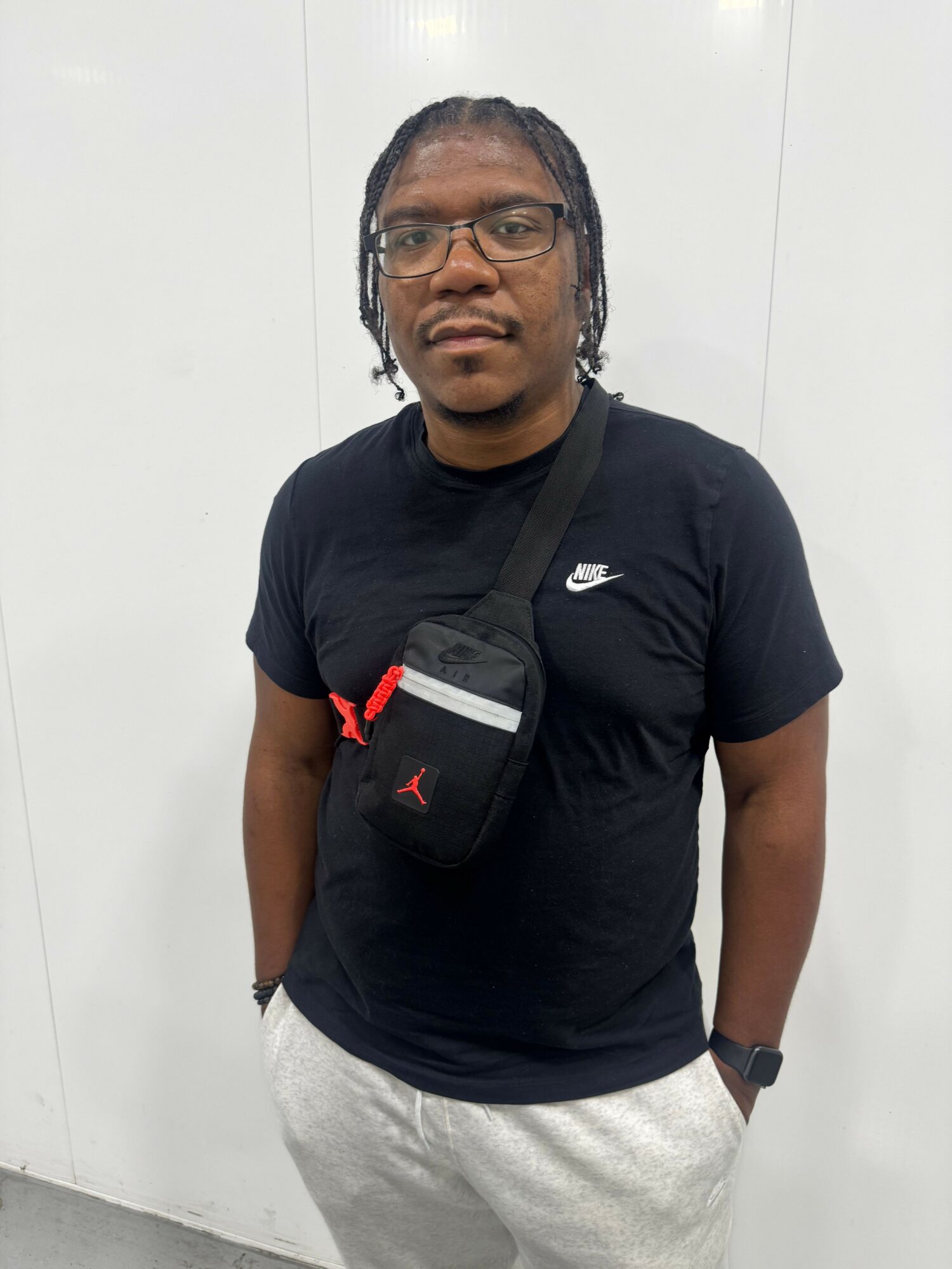 Person with glasses and braided hair standing against a plain white background, wearing a black t-shirt and light-colored pants.