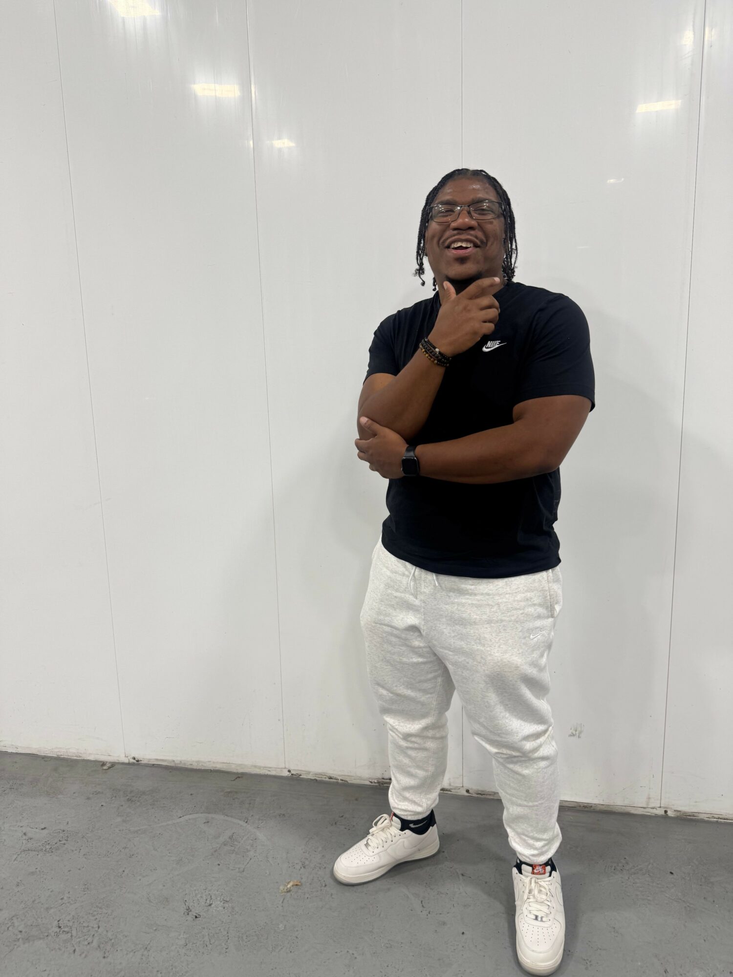 Person taking a mirror selfie, standing against a white wall, wearing black shirt and white pants, holding a phone to face.