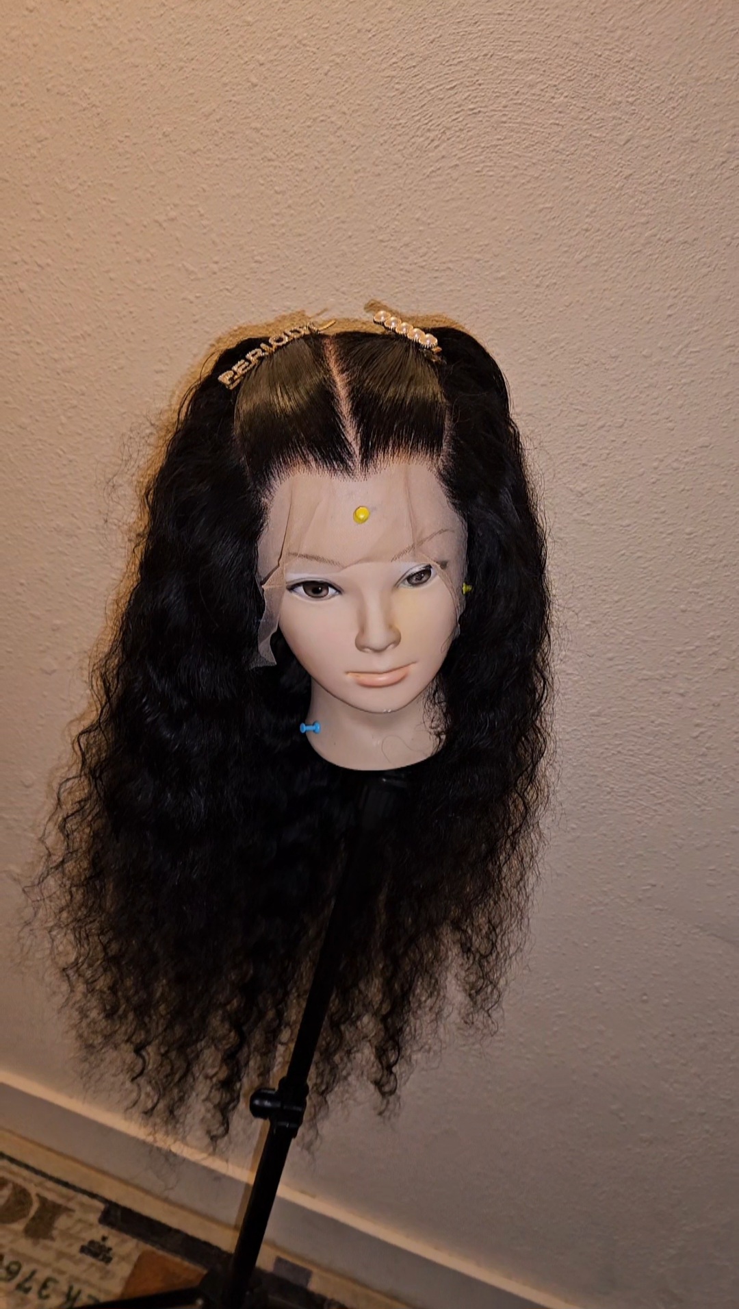 Doll head with long, curly black hair and a decorative headband, mounted on a stand against a plain wall.