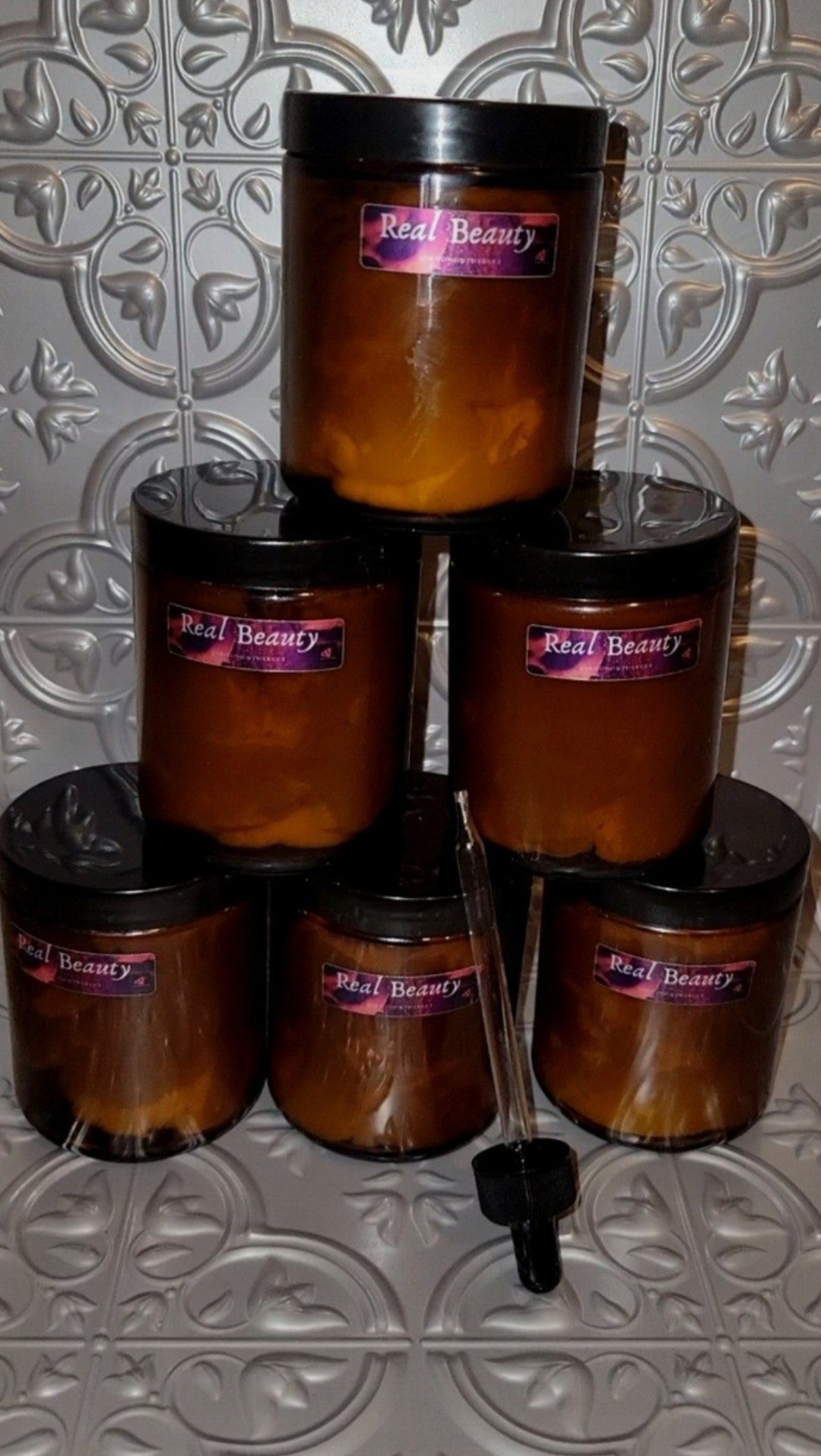 Six amber-colored jars with black lids arranged in a pyramid on a decorative tiled surface, labels reading 'Real Beauty'.