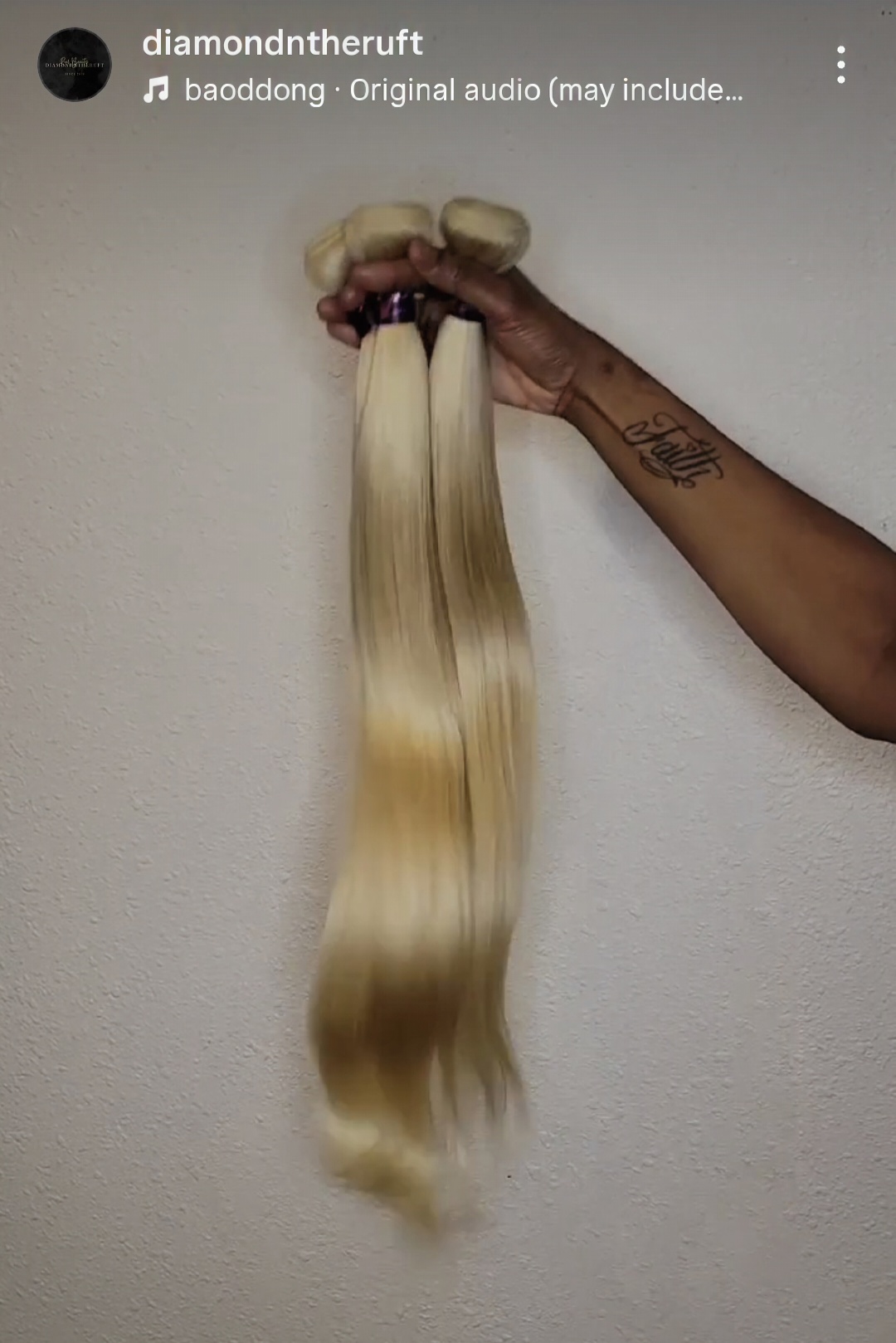 Person's arm holding long, straight blonde hair extension against a plain wall.