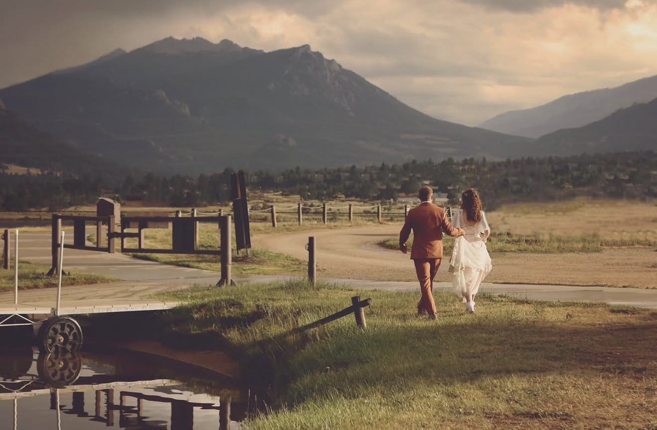 A couple walks hand in hand on a grassy path with mountains in the background, reflecting in a small pond.