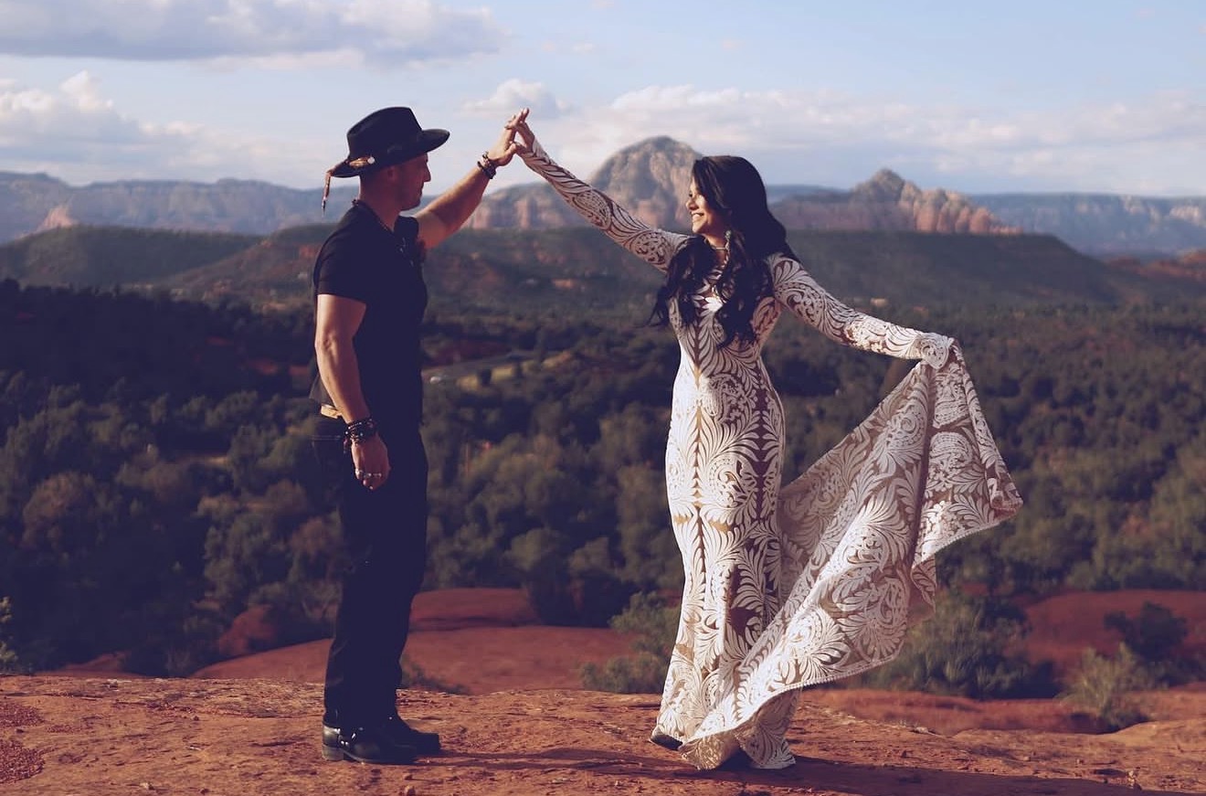 Two people dancing outdoors with mountains in the background, holding hands, one in a black hat and dress, the other in a flowing dress.