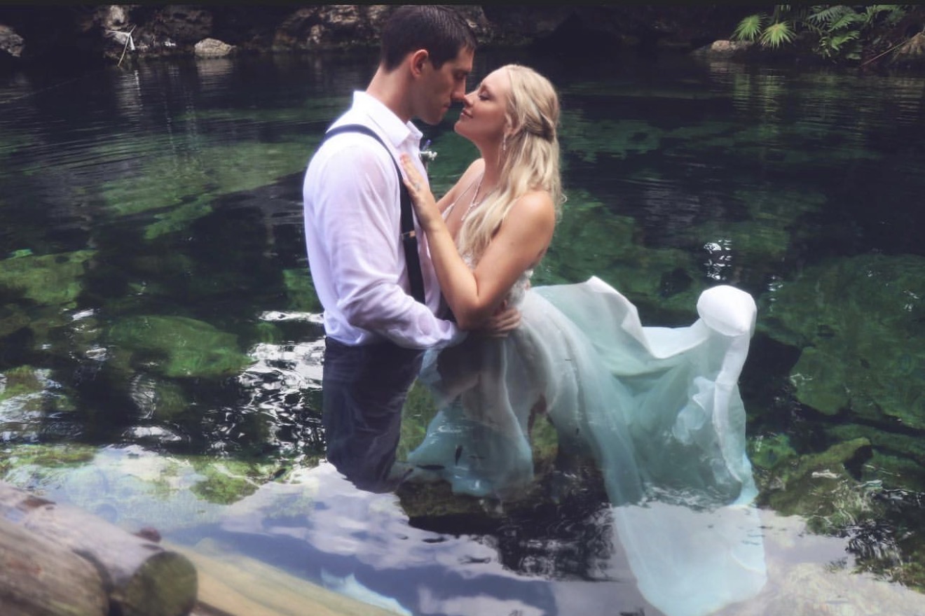 A couple stands in a body of water, embracing and about to kiss, with a natural outdoor background.