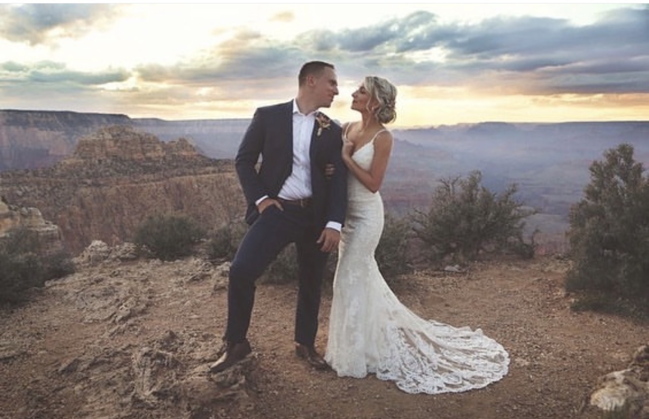 A couple in wedding attire standing on a rocky landscape with a canyon and sunset in the background.