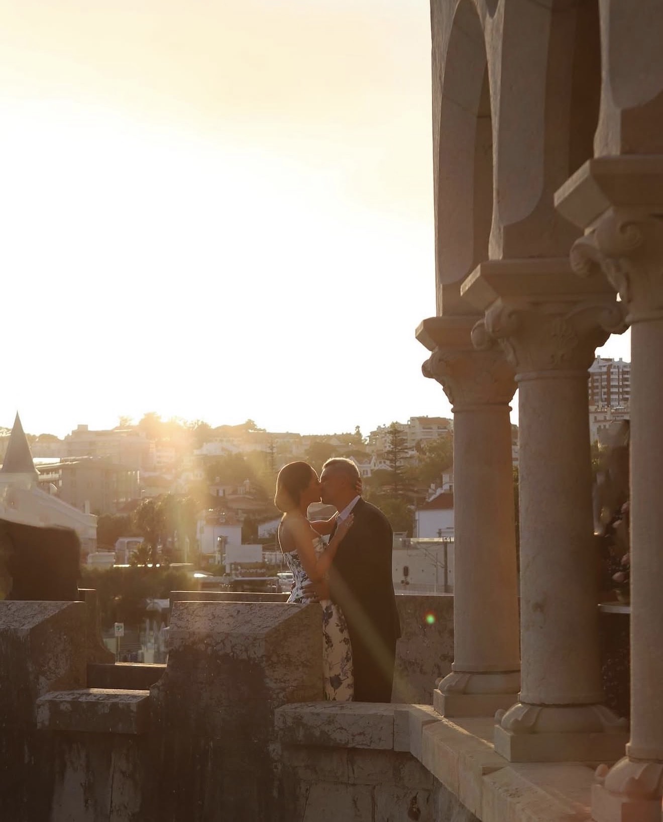 Two people dancing on a balcony with columns, sunset in the background, cityscape visible behind them.