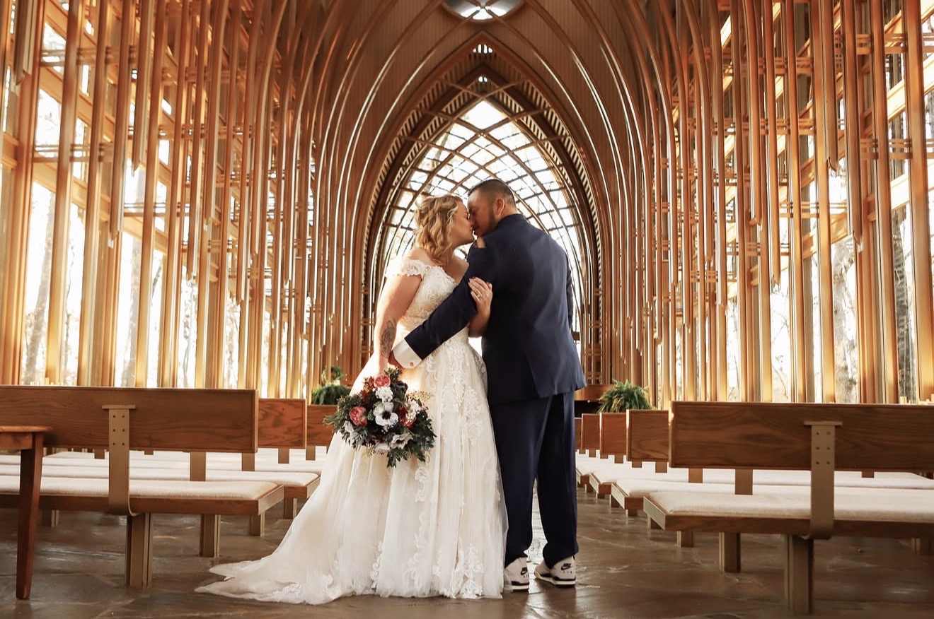 A bride and groom kiss inside a wooden church with arched ceiling and benches, holding a bouquet of flowers.
