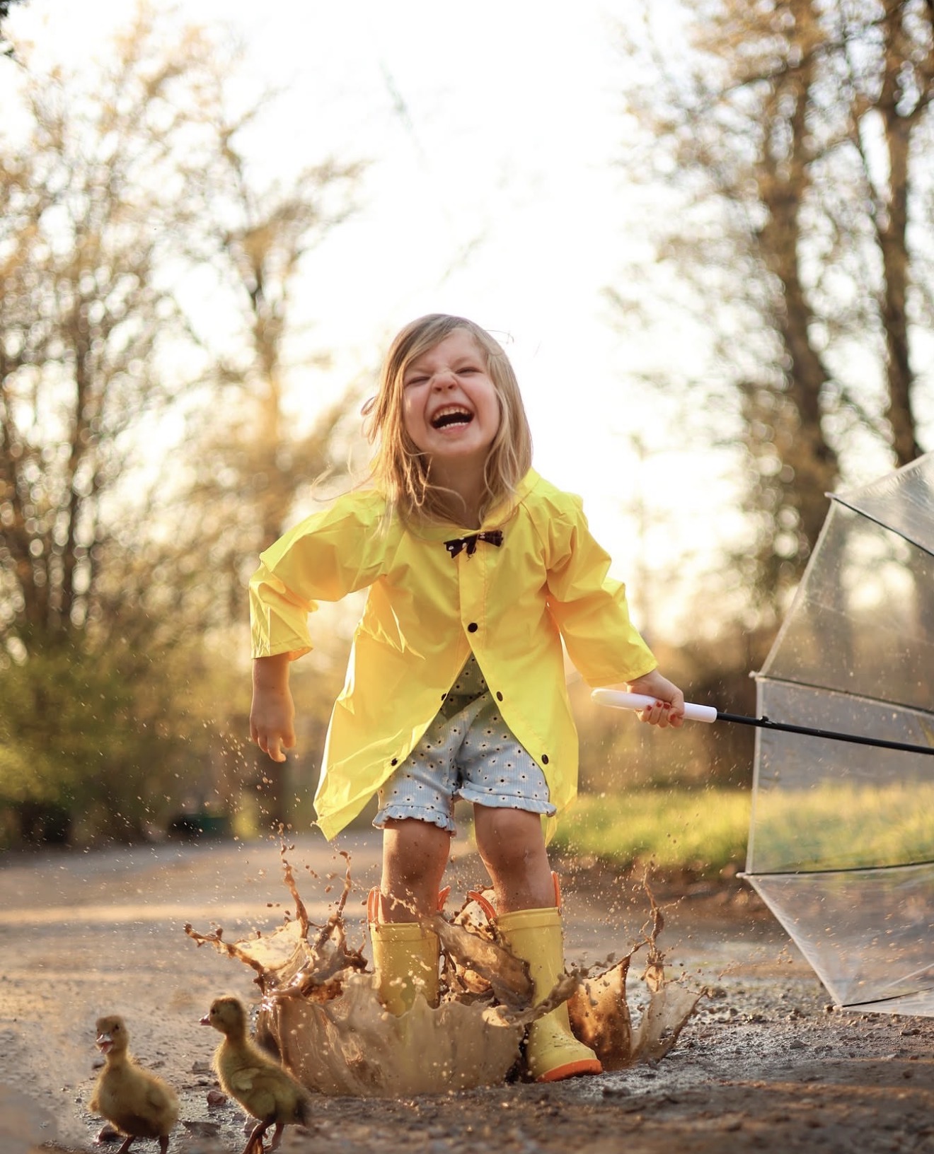 Child in yellow raincoat and rain boots splashing in puddle outdoors, smiling and holding an umbrella, trees in background
