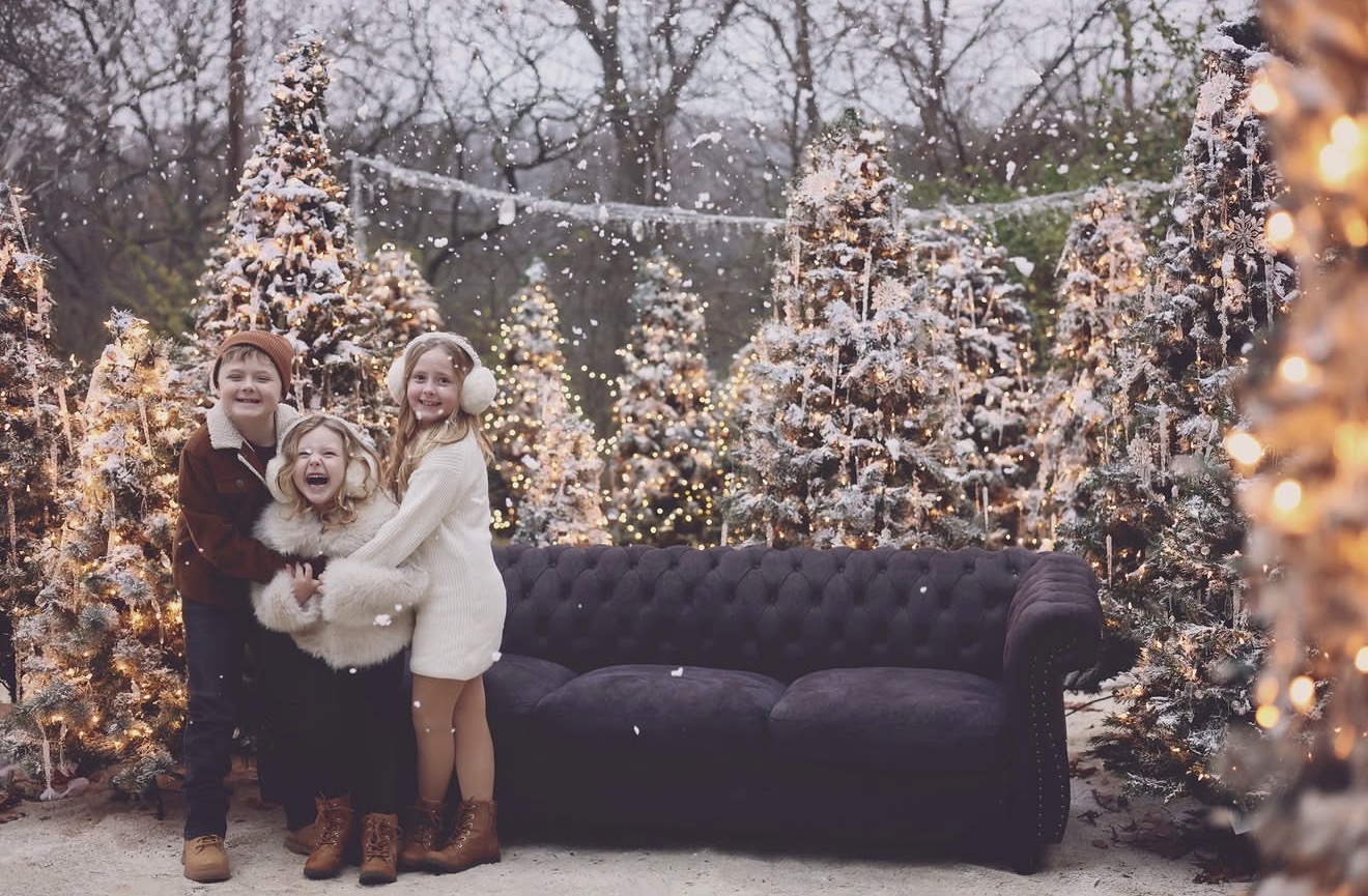 Four children stand outdoors in a snowy scene with decorated trees and string lights, smiling and hugging each other, with a black sofa nearby.