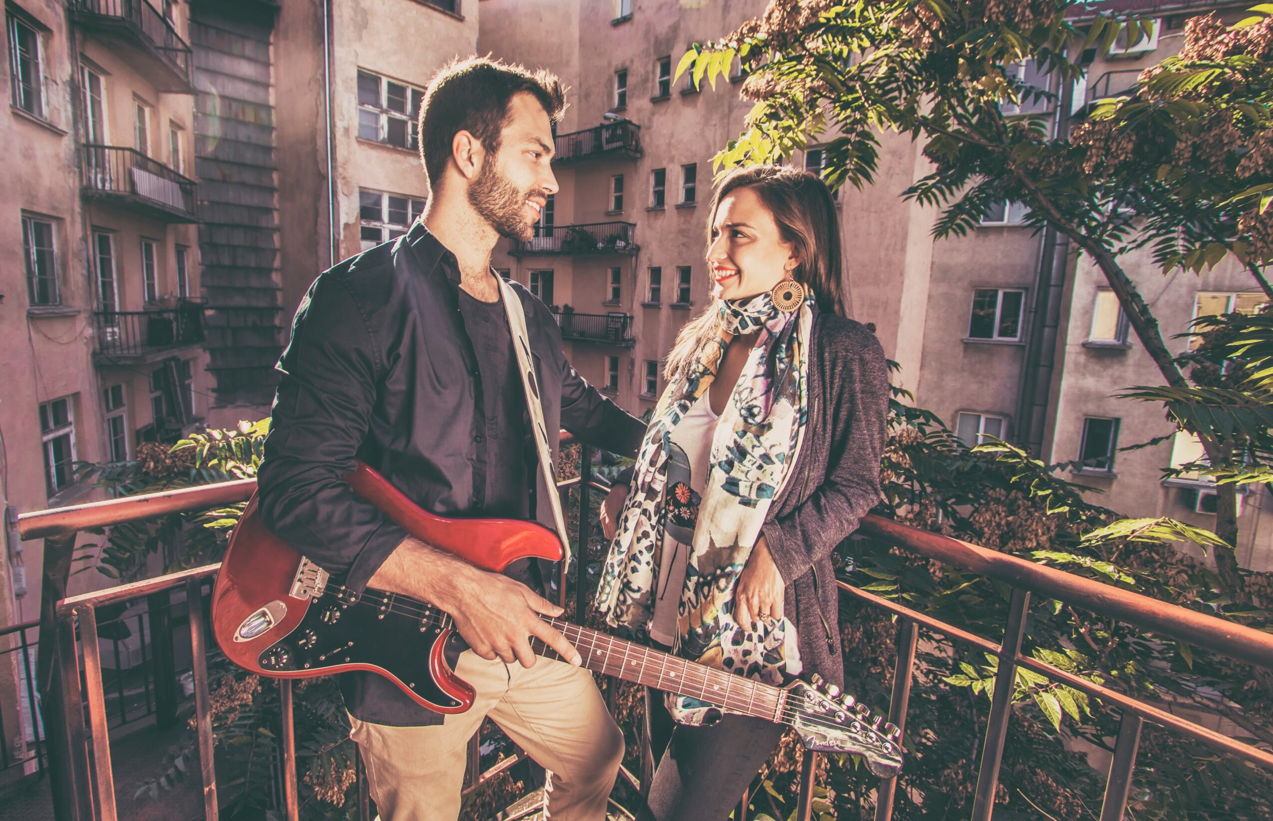 A man and woman stand on a balcony with buildings in the background, talking and smiling, with a guitar.