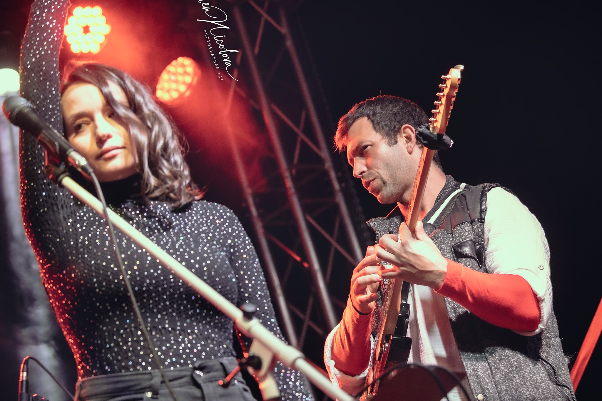 A woman and a man perform on stage with stage lights and equipment visible, woman holding a microphone, man playing guitar.