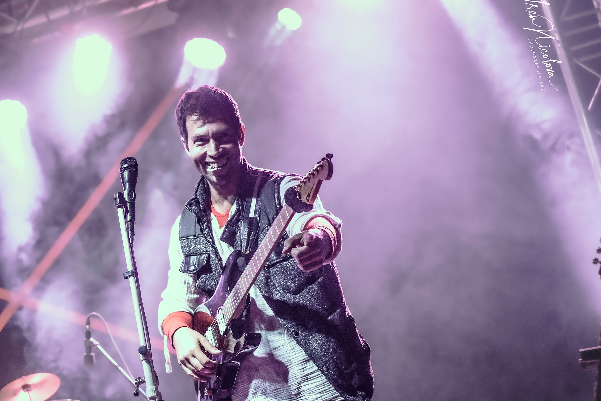 Person on stage holding a guitar, smiling, with stage lights and fog in the background.