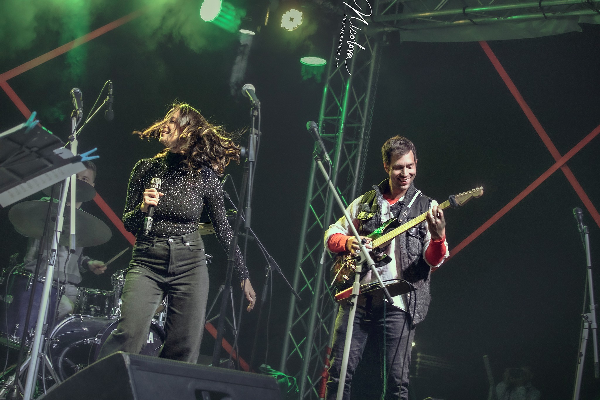 Two musicians perform on stage, one singing and the other playing guitar, with stage lights and equipment visible.
