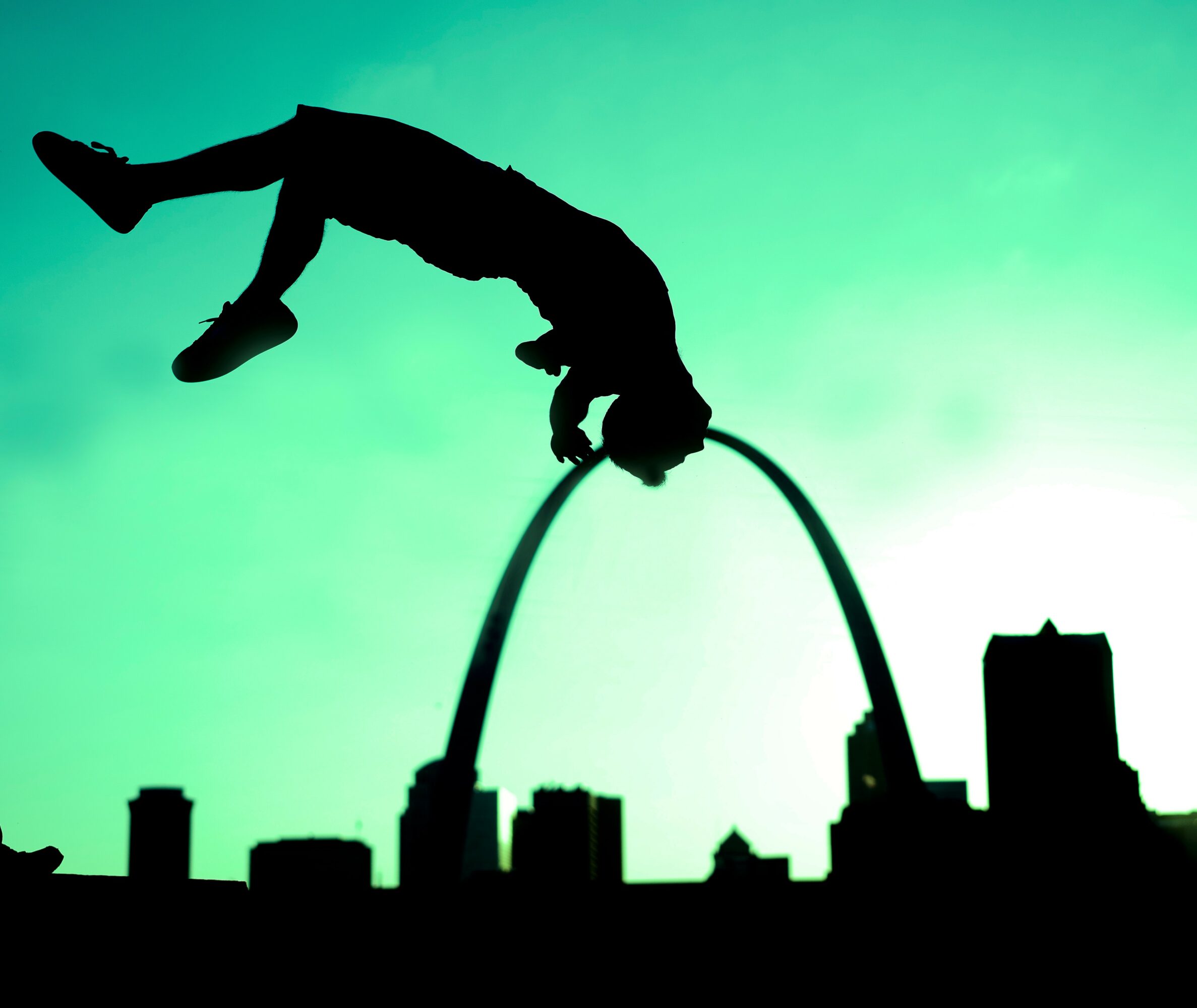 Silhouette of a person performing a flip on a curved structure against a green sky, city skyline below.