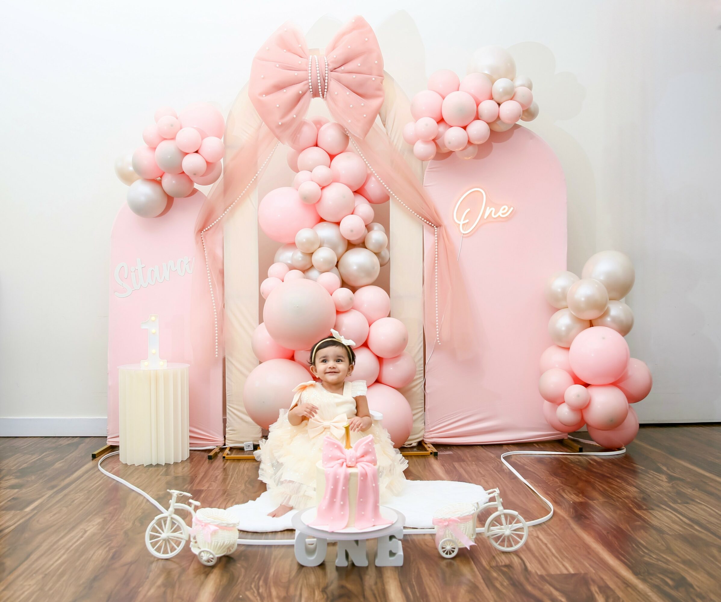 Child in a white dress with a pink cake, birthday decorations, balloons, and a pink backdrop with a large bow.