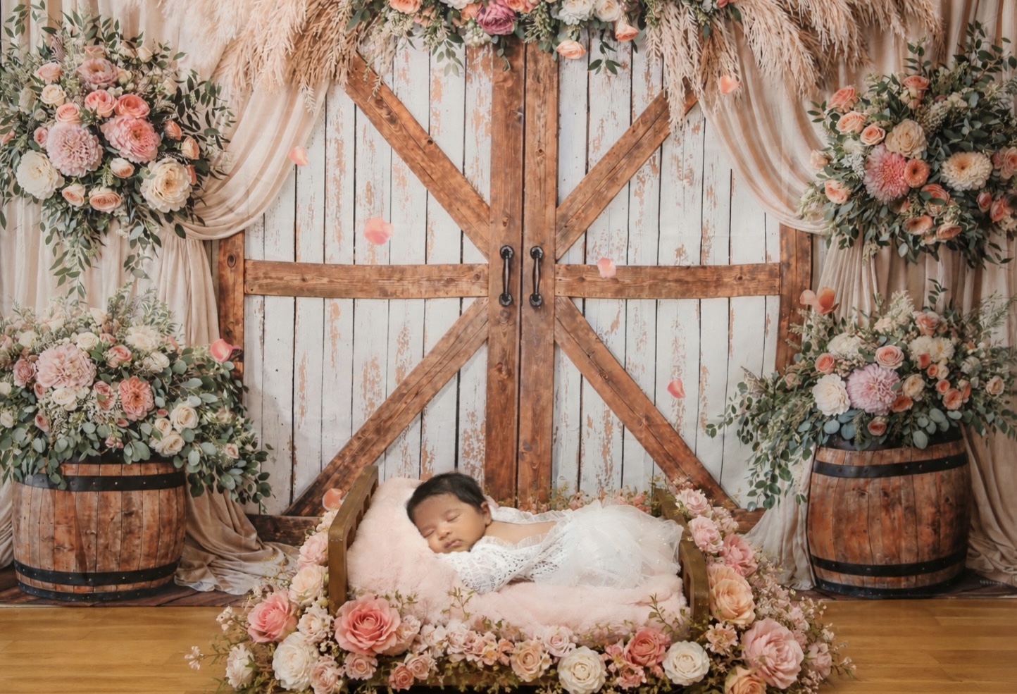 Baby lying on a floral bed in front of a wooden barn door with flower arrangements on barrels.