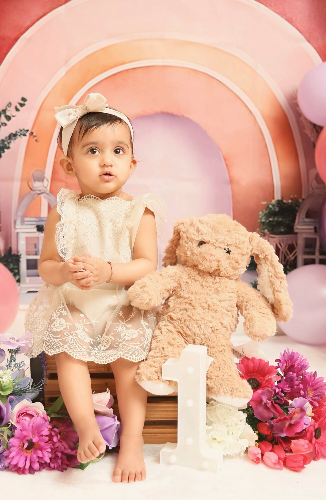 Young girl in a cream dress with a bow headband sitting next to a teddy bear, surrounded by pink and purple decorations and flowers.