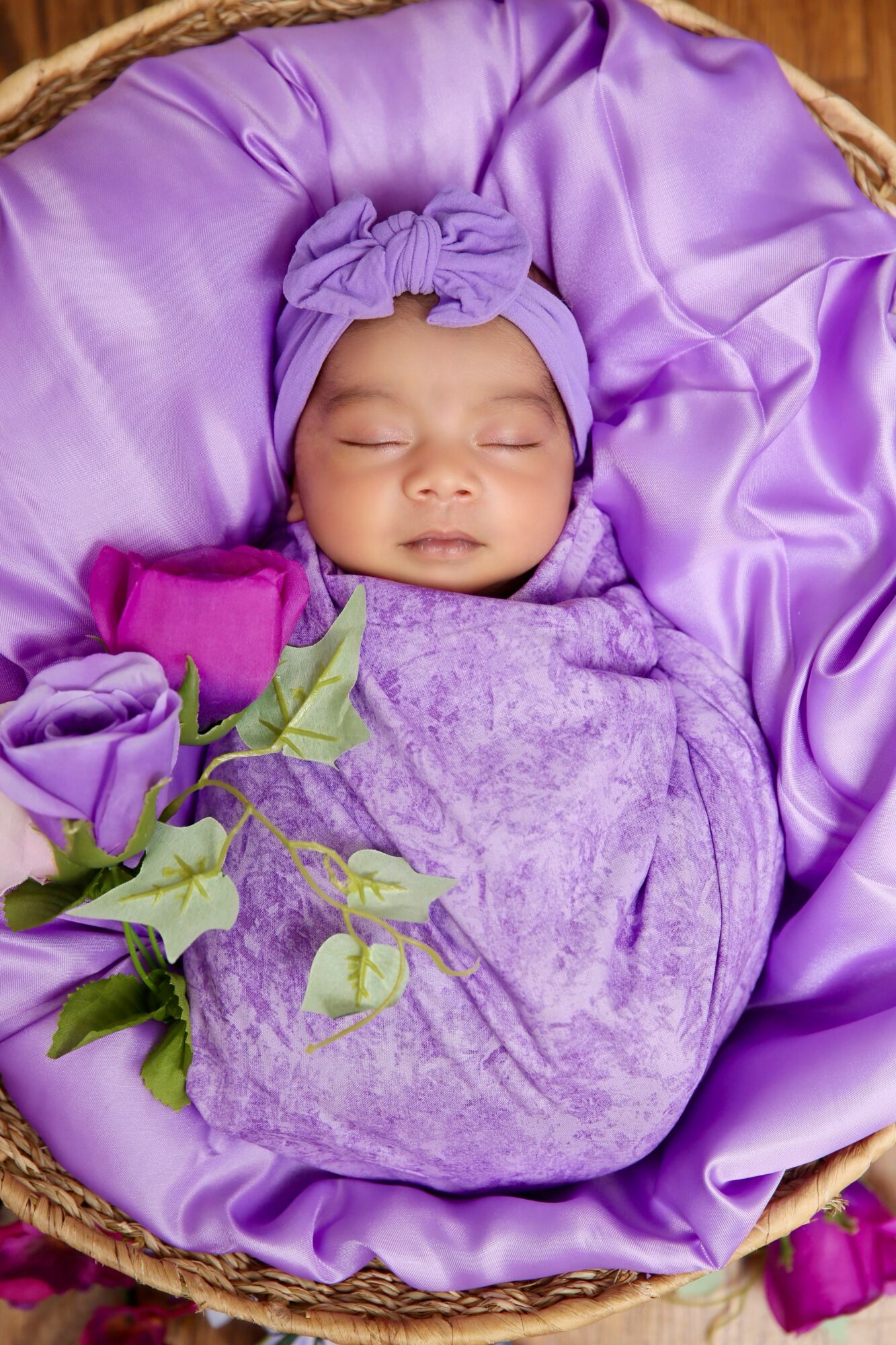 Baby wrapped in purple cloth, lying on purple satin fabric in a basket, with purple flowers nearby.