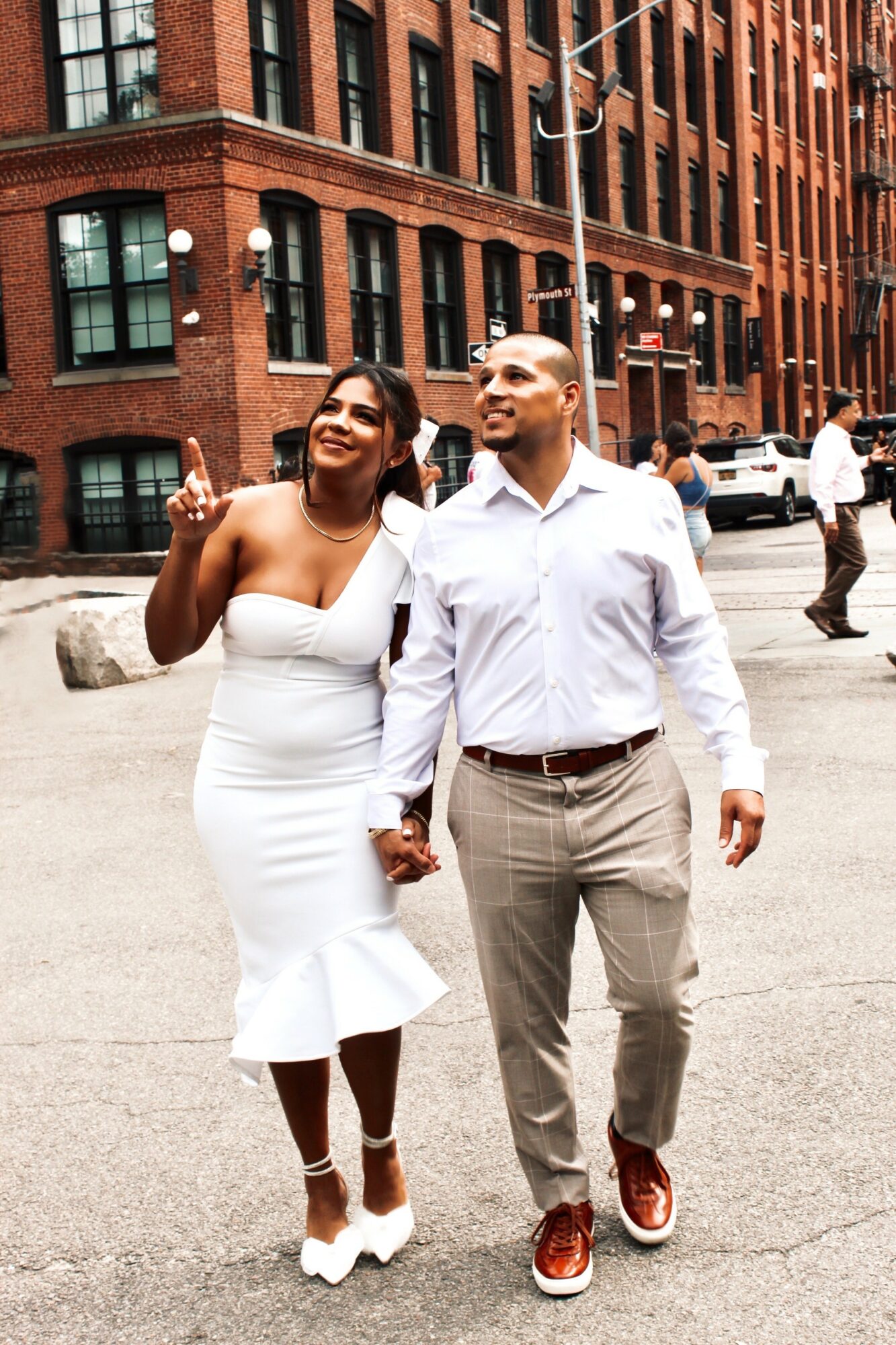 A couple walking hand in hand on a city street, smiling, with brick buildings in the background.