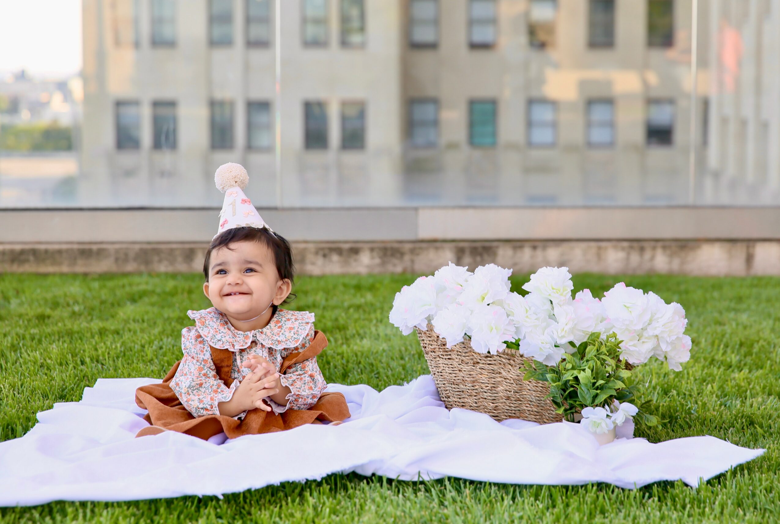 Baby girl sitting on white cloth outdoors with a basket of white flowers, smiling, in front of a building.