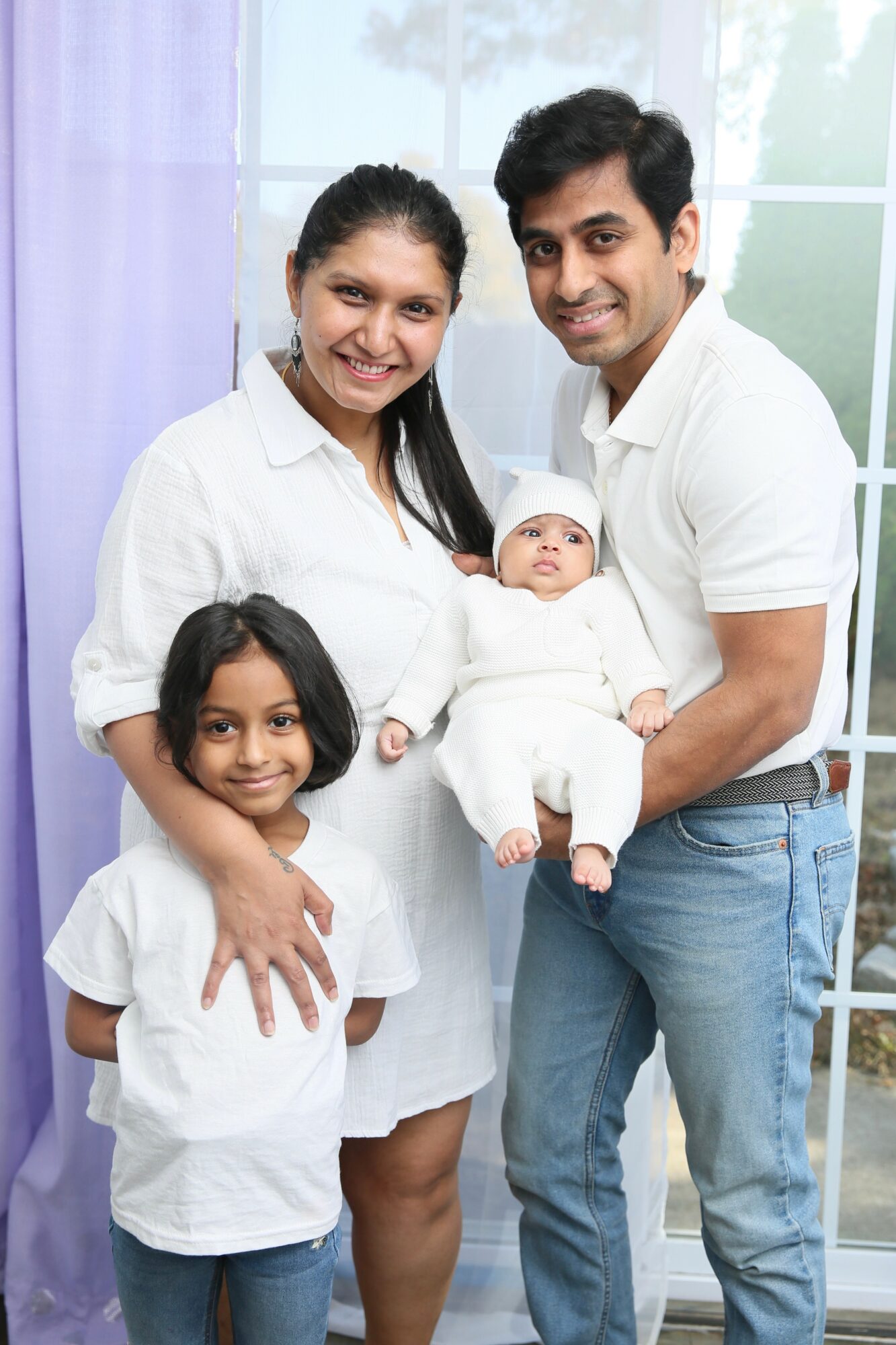 Family of four with a baby, standing indoors near a window, smiling, with light-colored clothing.