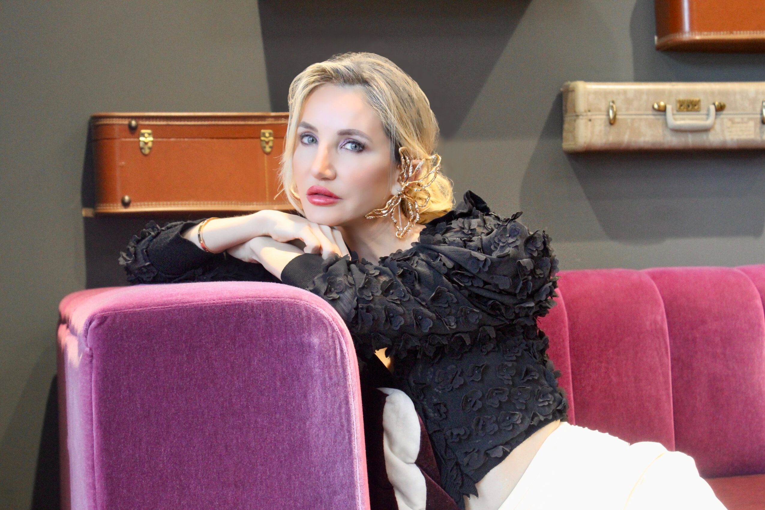 Woman with blonde hair resting her head on her arms on a pink sofa, wearing a black lace top, in a room with vintage suitcases on the wall.
