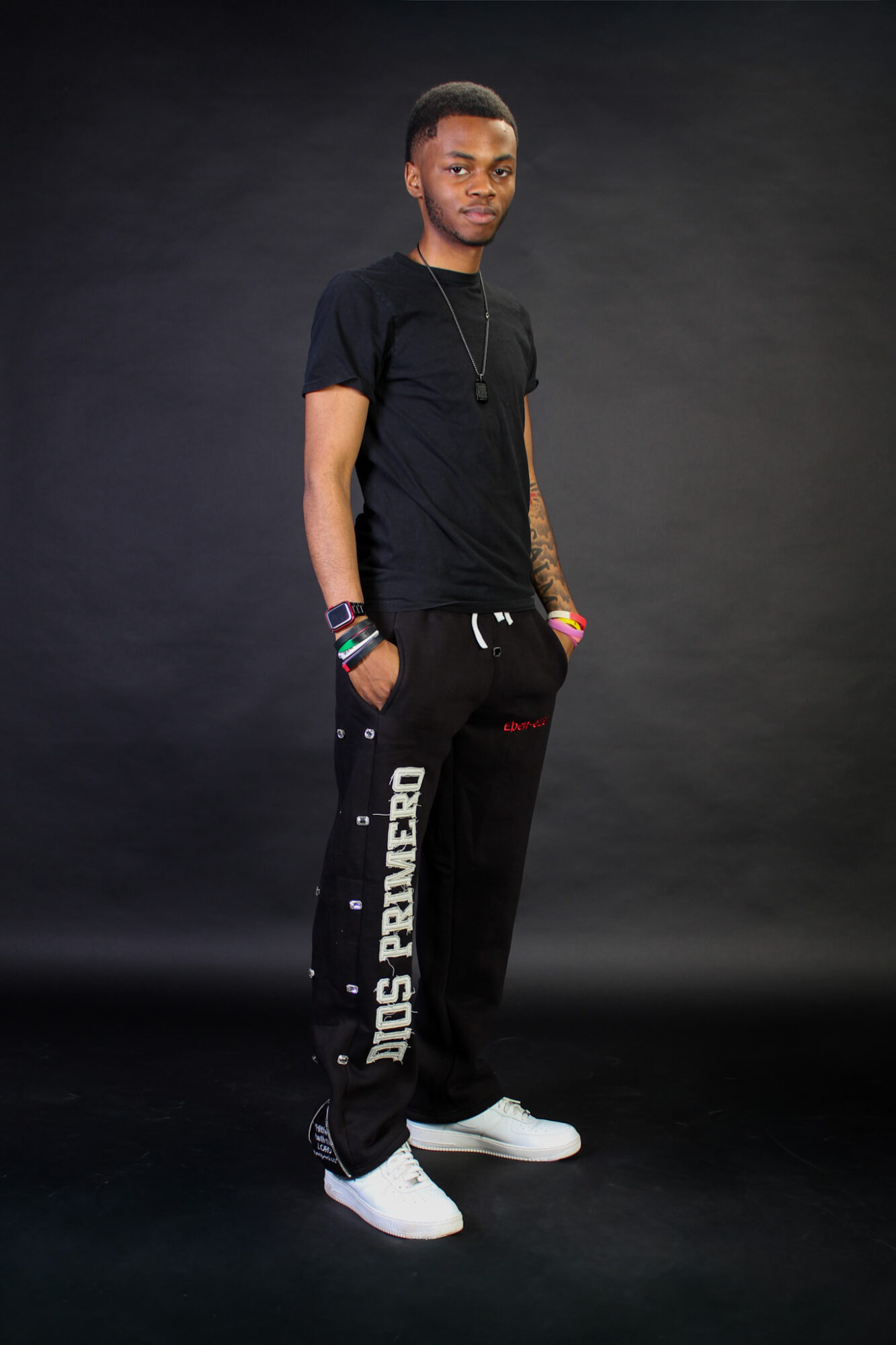 Young man standing against a dark background, wearing black t-shirt, black sweatpants, and white sneakers, with colorful bracelets.