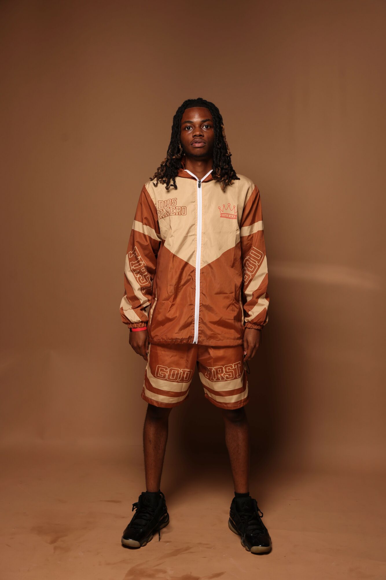 Person standing against a plain brown wall, wearing a patterned jacket, shorts, and sneakers, with long curly hair.