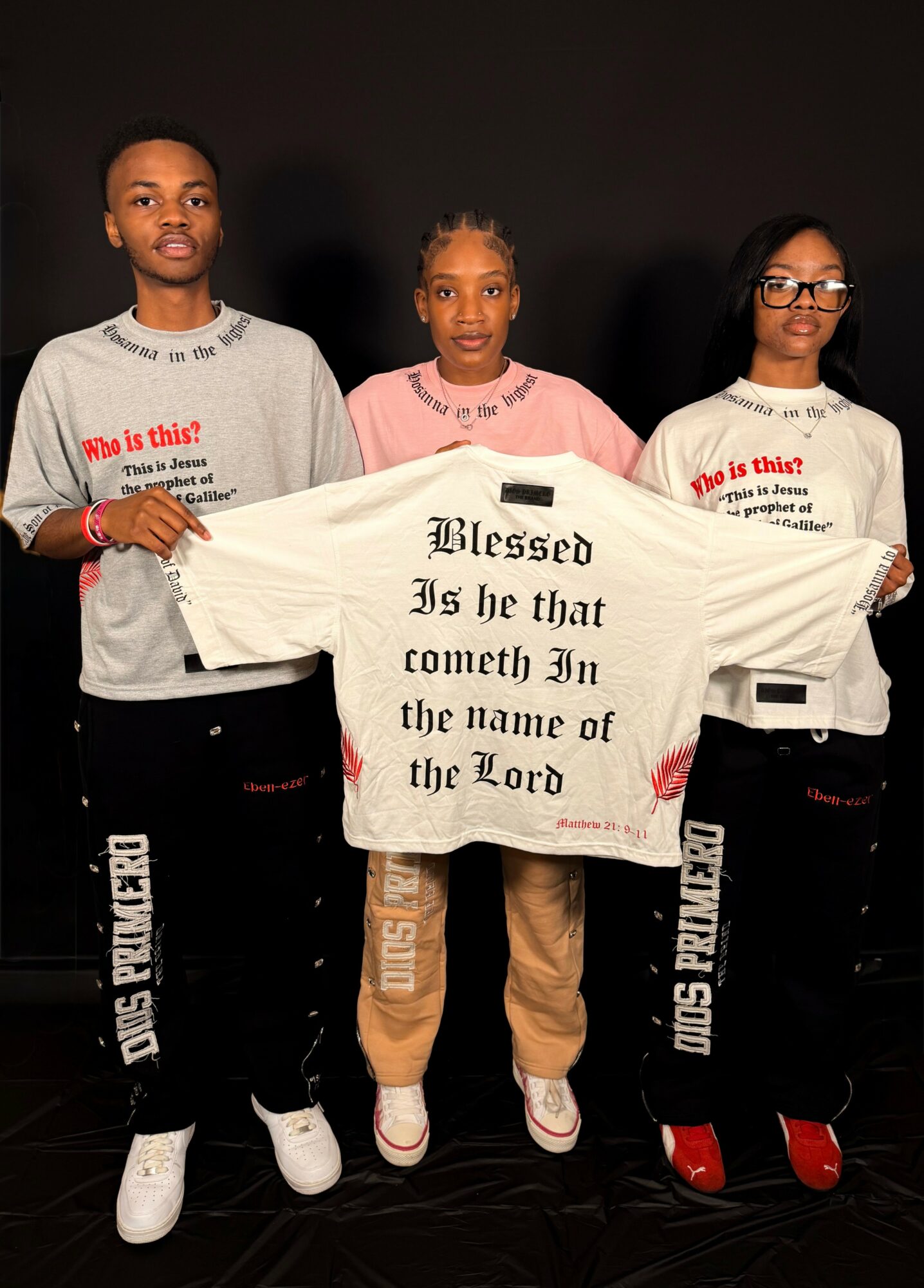 Three people holding a white shirt with black text, standing against a dark background. Two wear glasses, one has braided hair.