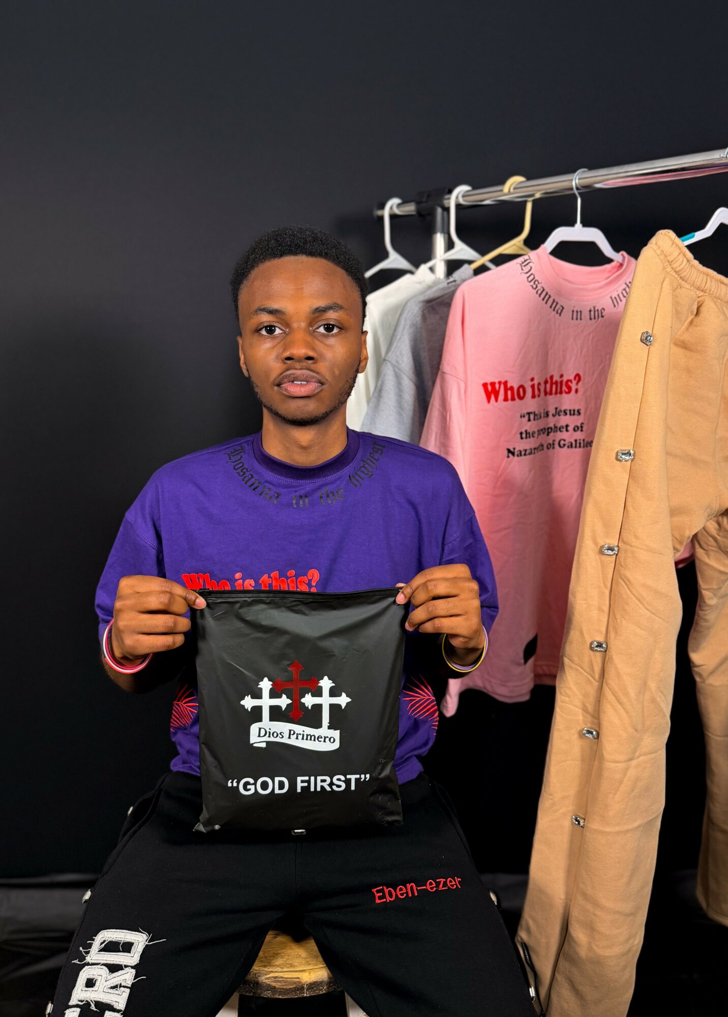 Young man holding a black bag with white text, standing next to clothing on a rack against a dark background.