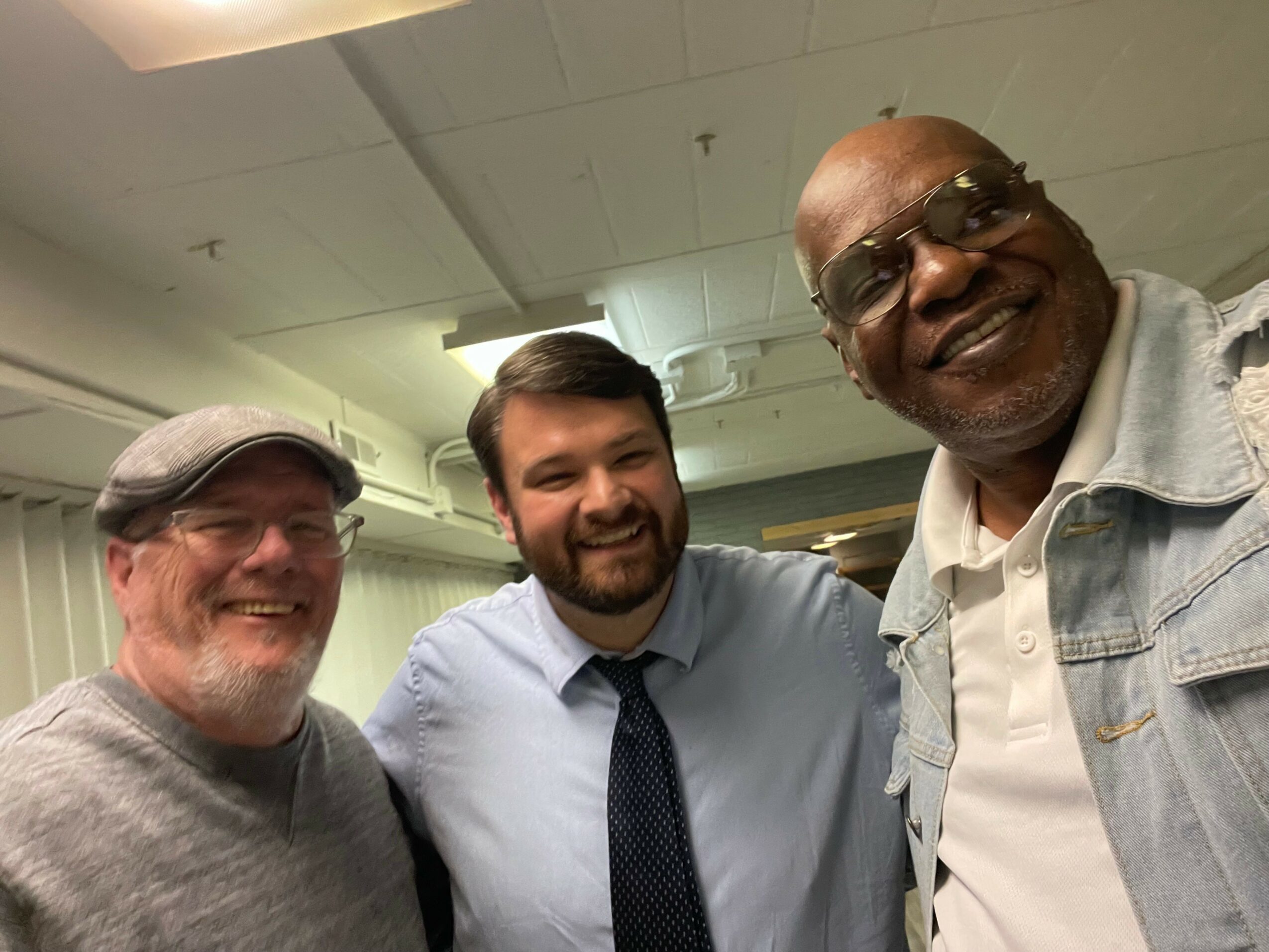 Three men smiling indoors, one wearing glasses and a cap, another with a beard, and the third with sunglasses.
