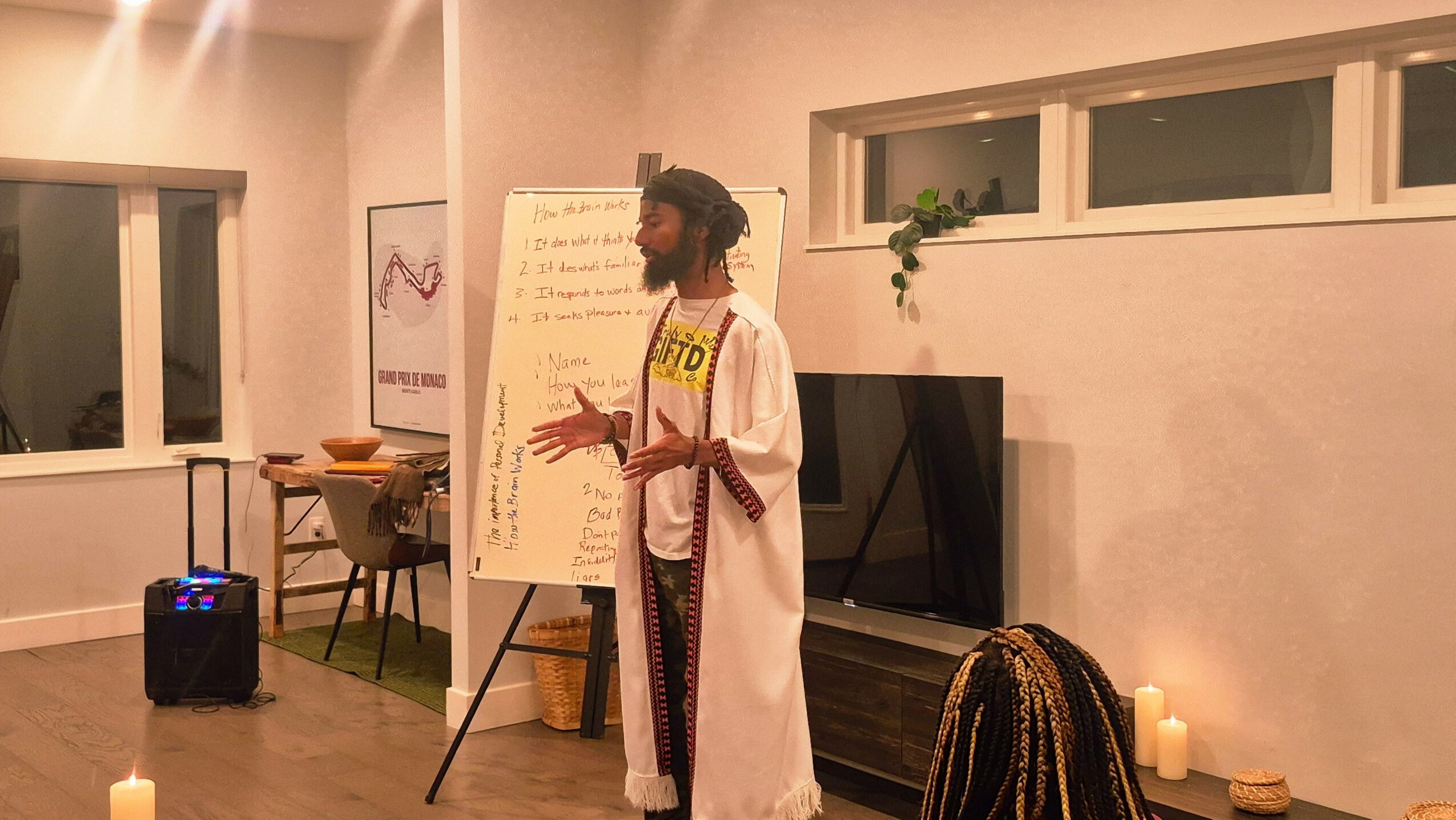Person standing in front of a whiteboard, speaking or presenting, in a room with candles and a window.