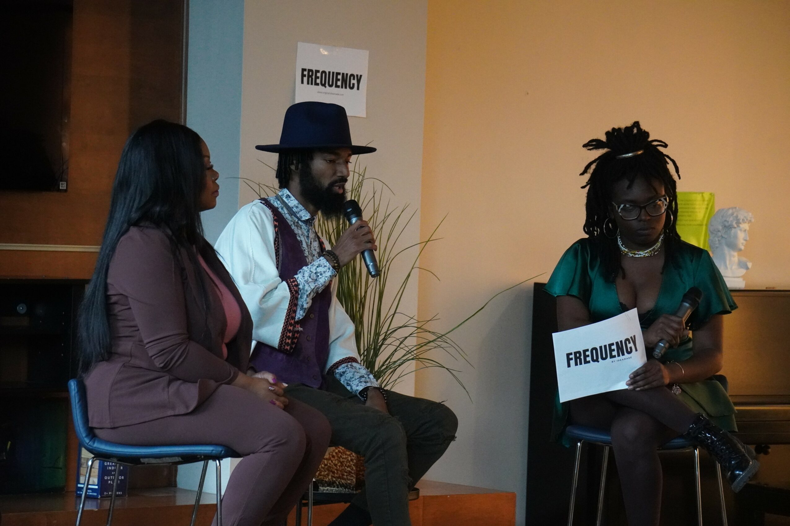 Three people sitting on chairs, two women and one man, with signs reading 'FREQUENCY' in front of a beige wall.