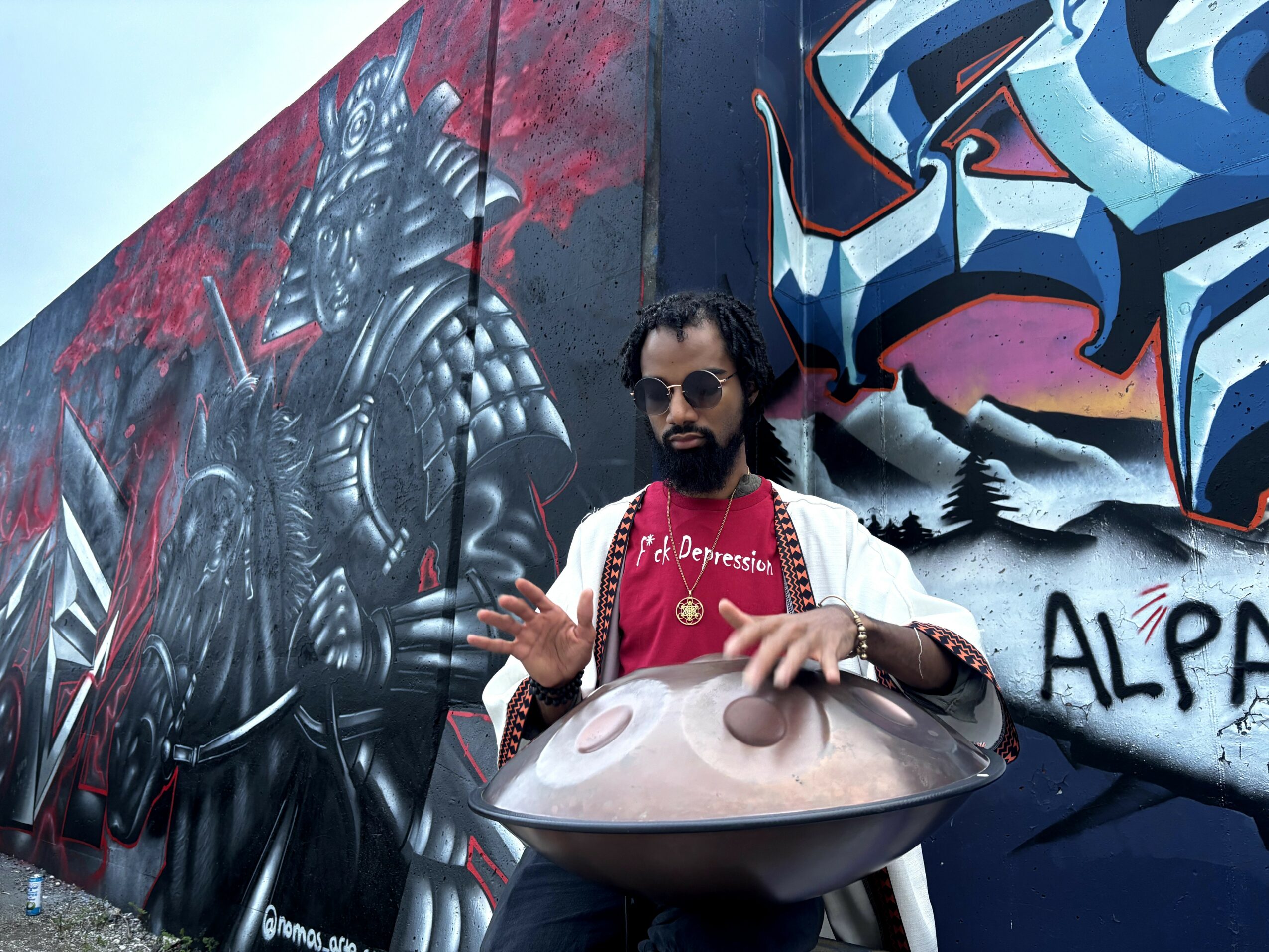 Man with sunglasses and beard playing a handpan instrument in front of graffiti wall with a knight and abstract art.