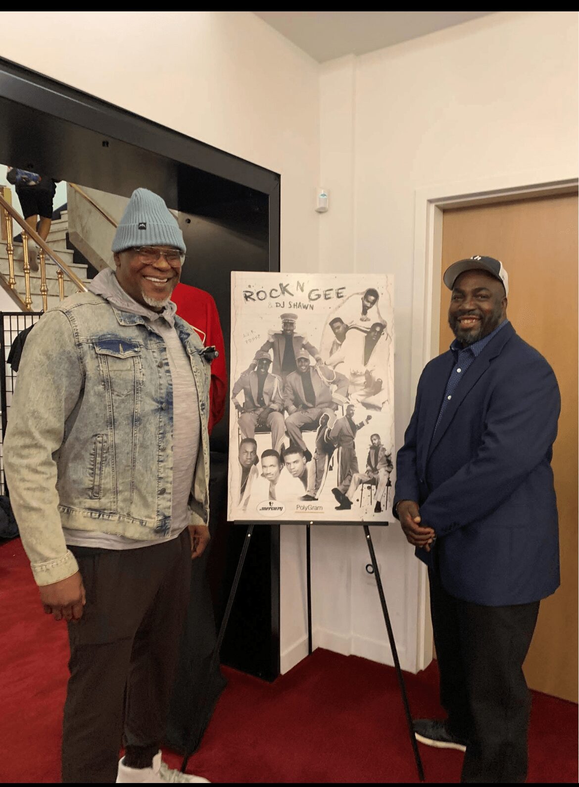 Two men standing next to a poster on an easel in a room with white walls and a door, smiling at the camera.